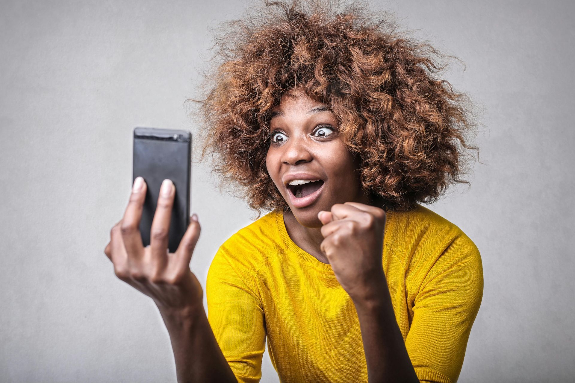 A woman in a yellow shirt is holding a cell phone in her hand and acting surprised. 