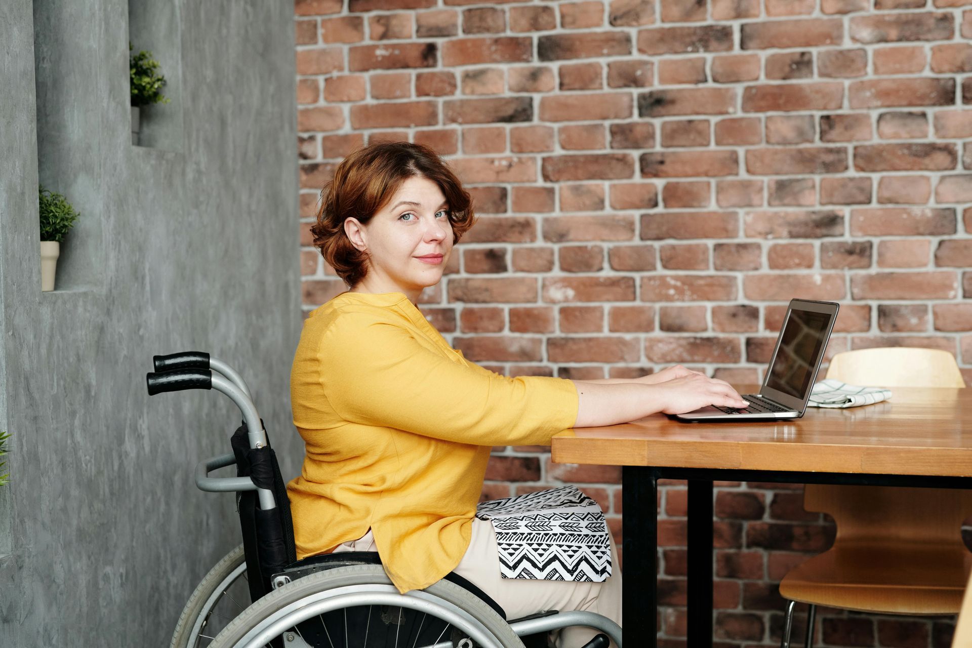 Woman in wheelchair working on laptop at a wooden table, brick wall background.