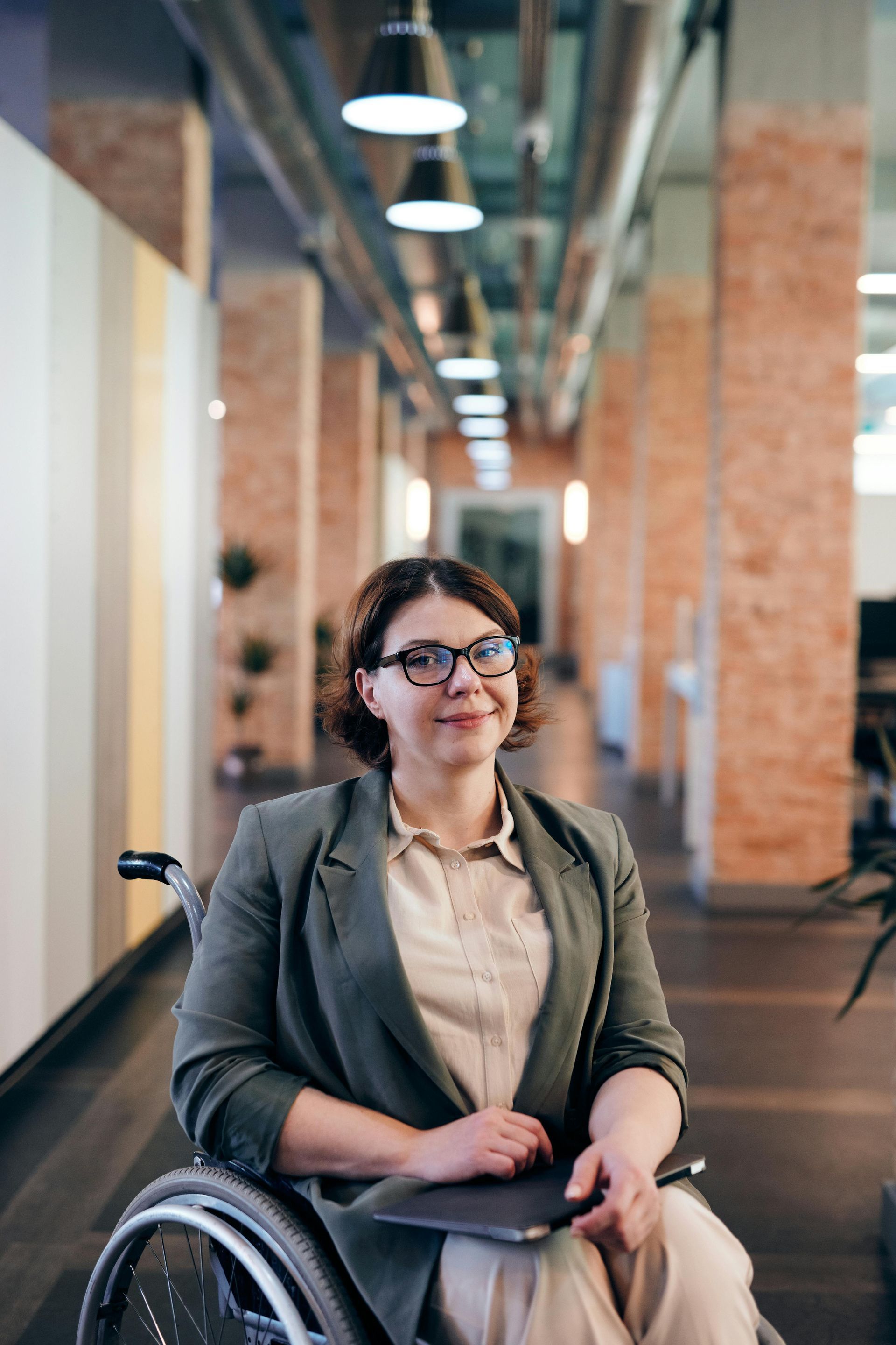 Woman in wheelchair in office hallway, holding tablet, wearing glasses, smiling.