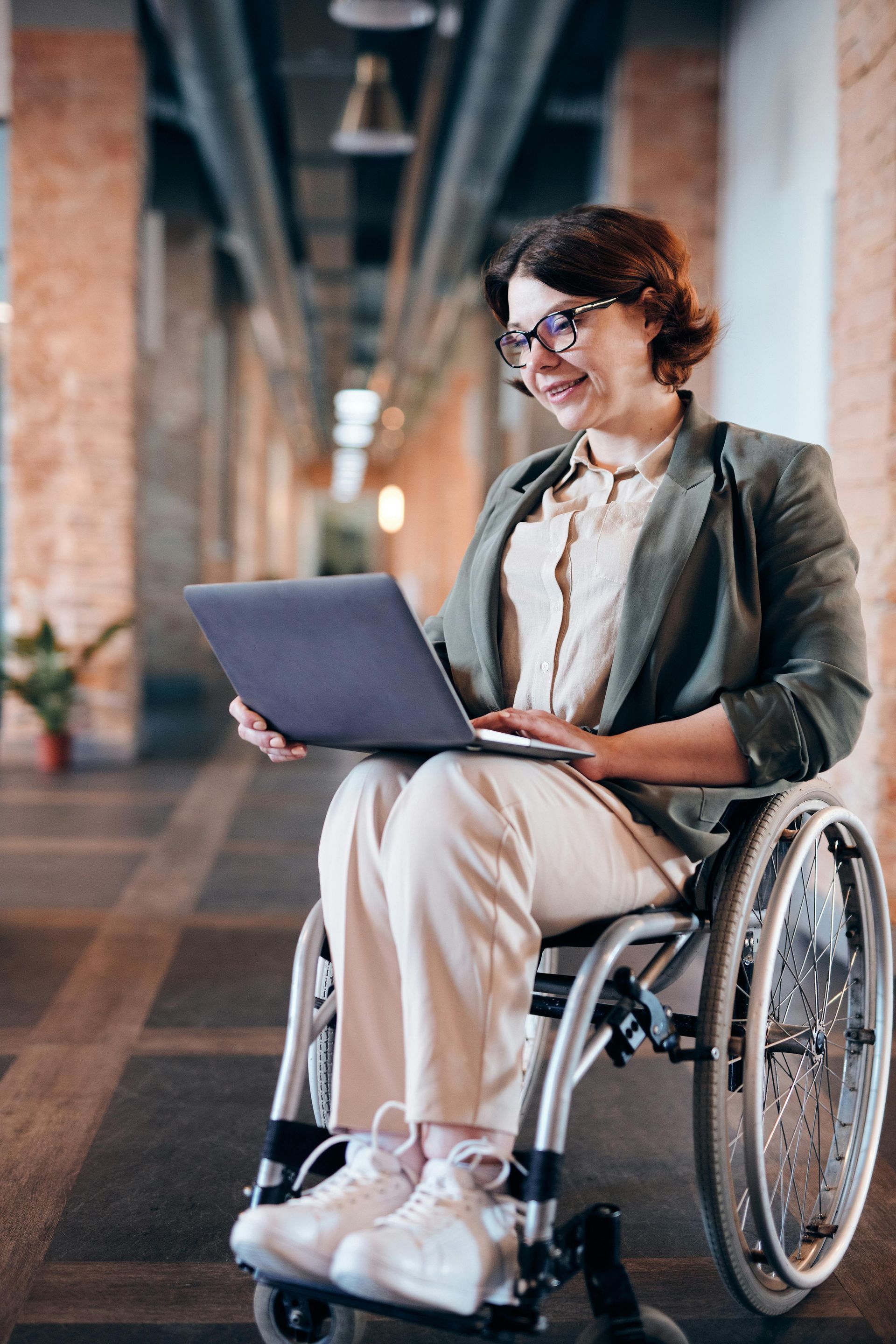 Woman in a wheelchair, working on a laptop, smiling, in a well-lit hallway.