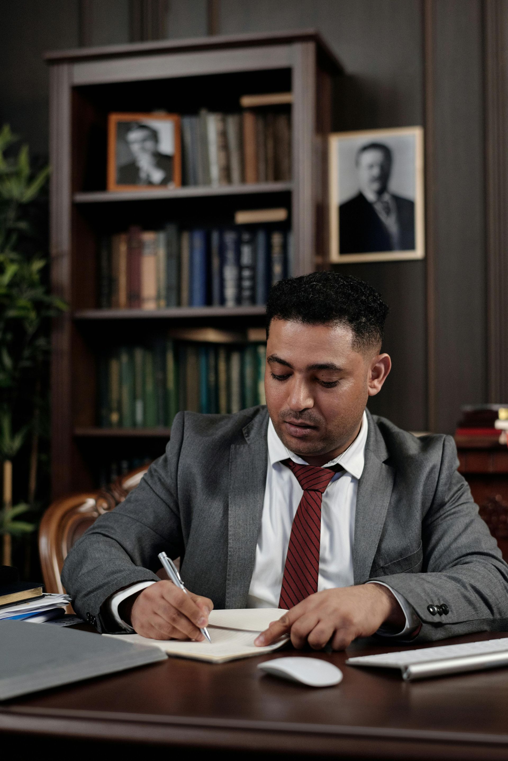 Man in gray suit writes in a notebook at a desk in an office with bookshelves and portraits.