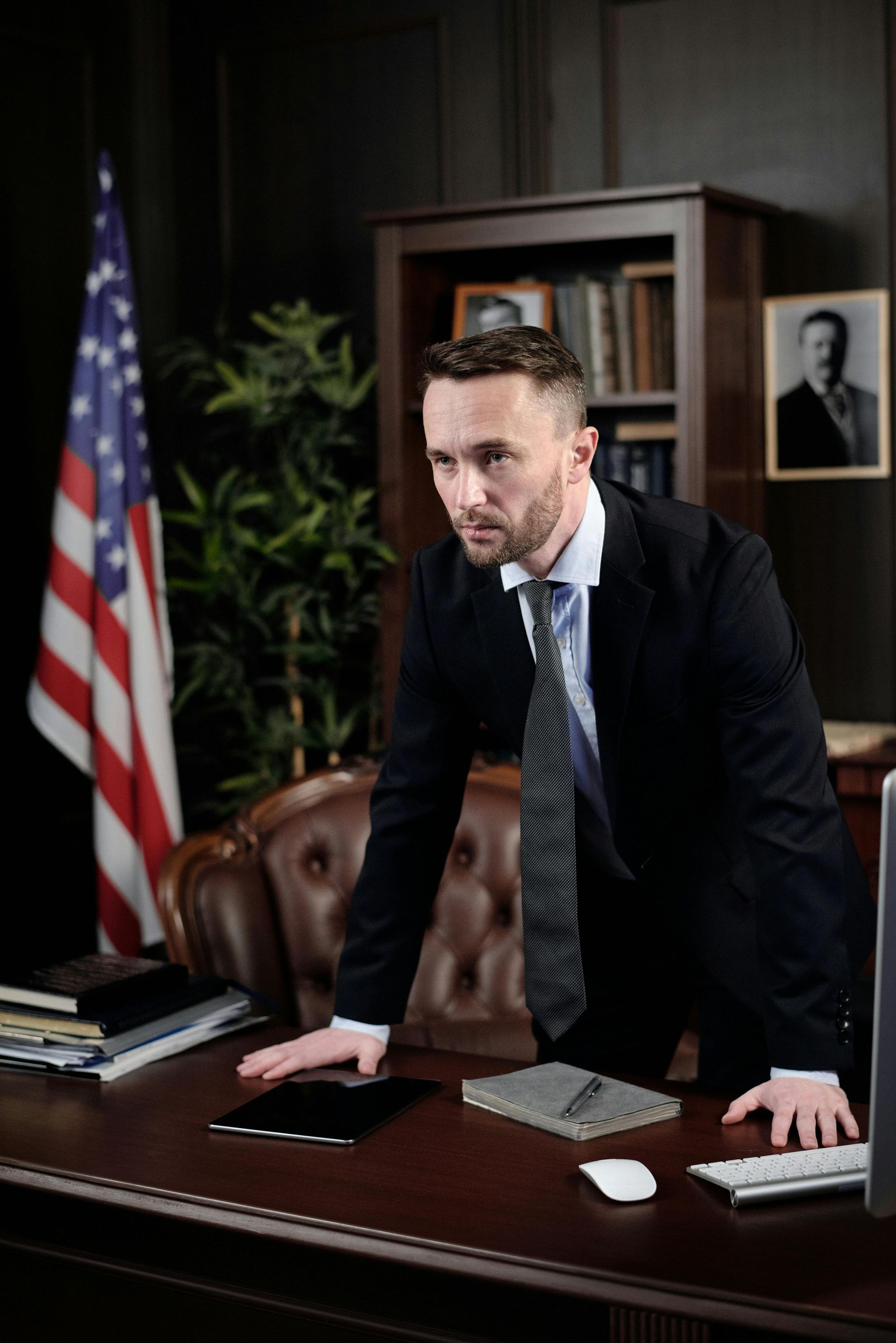 Man in a suit leans on a desk in an office with an American flag and bookshelf.