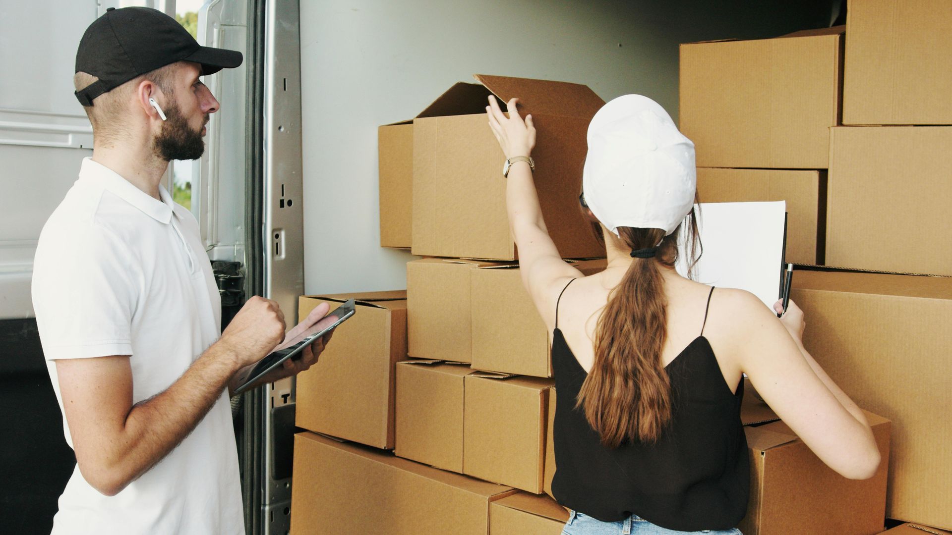 A delivery worker and a person in a black tank top stand inside a truck, checking and organizing cardboard boxes.