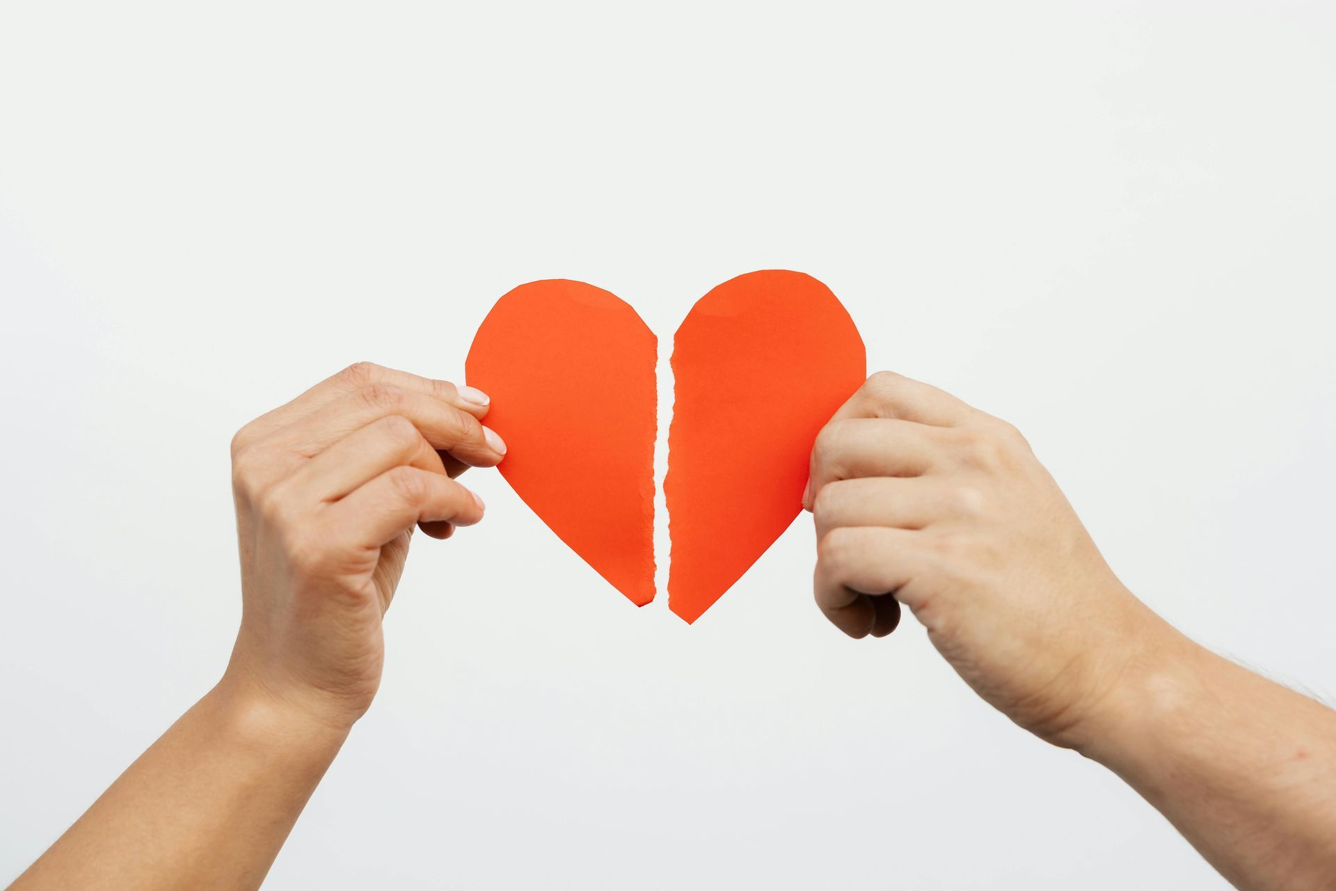 Two hands holding the two separate halves of a red paper heart against a plain white background.