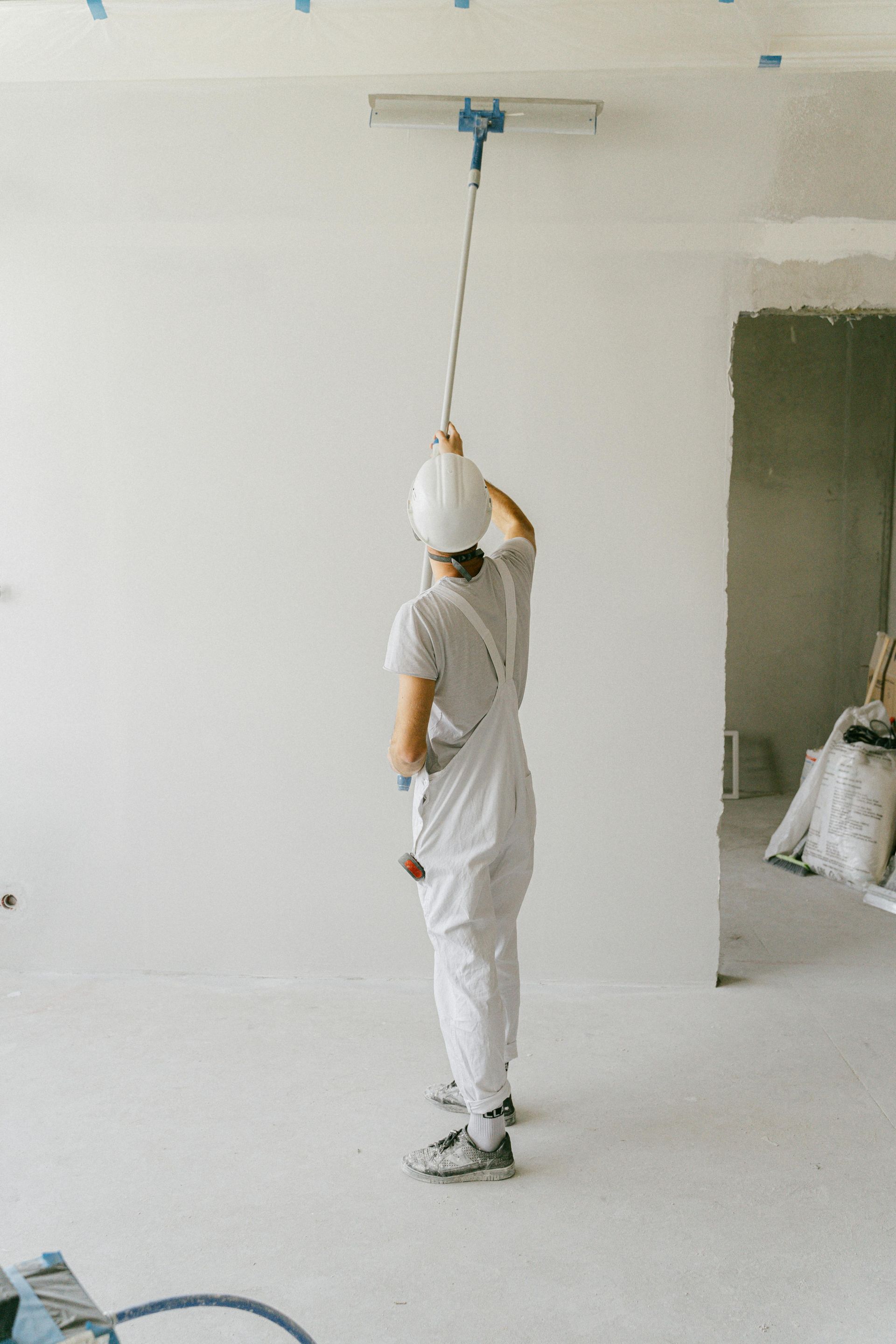 Person in white overalls and hard hat uses a long-handled tool to smooth plaster on a ceiling.