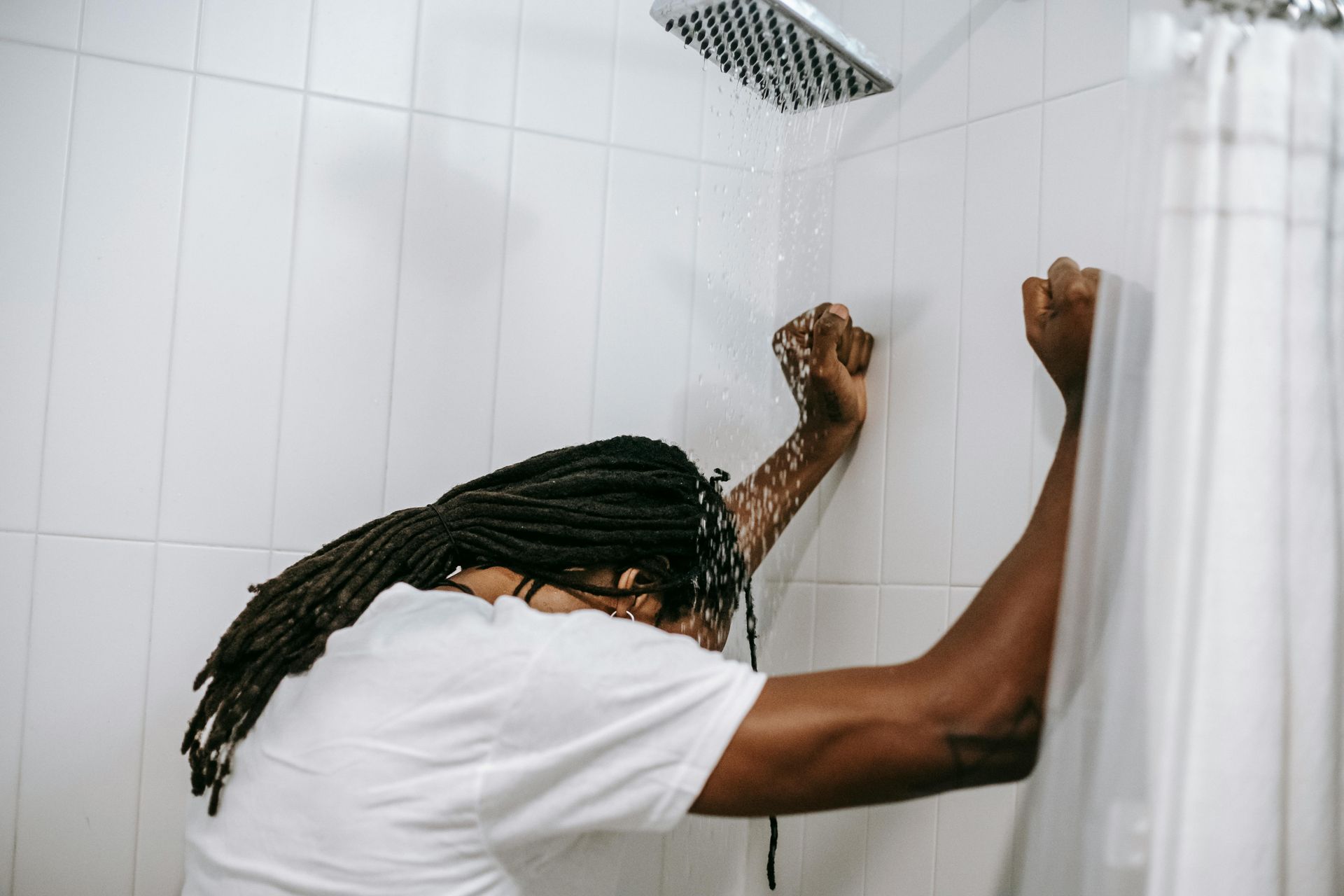 Person in a white t-shirt stands in a shower, head down, arms raised against the tiled wall.