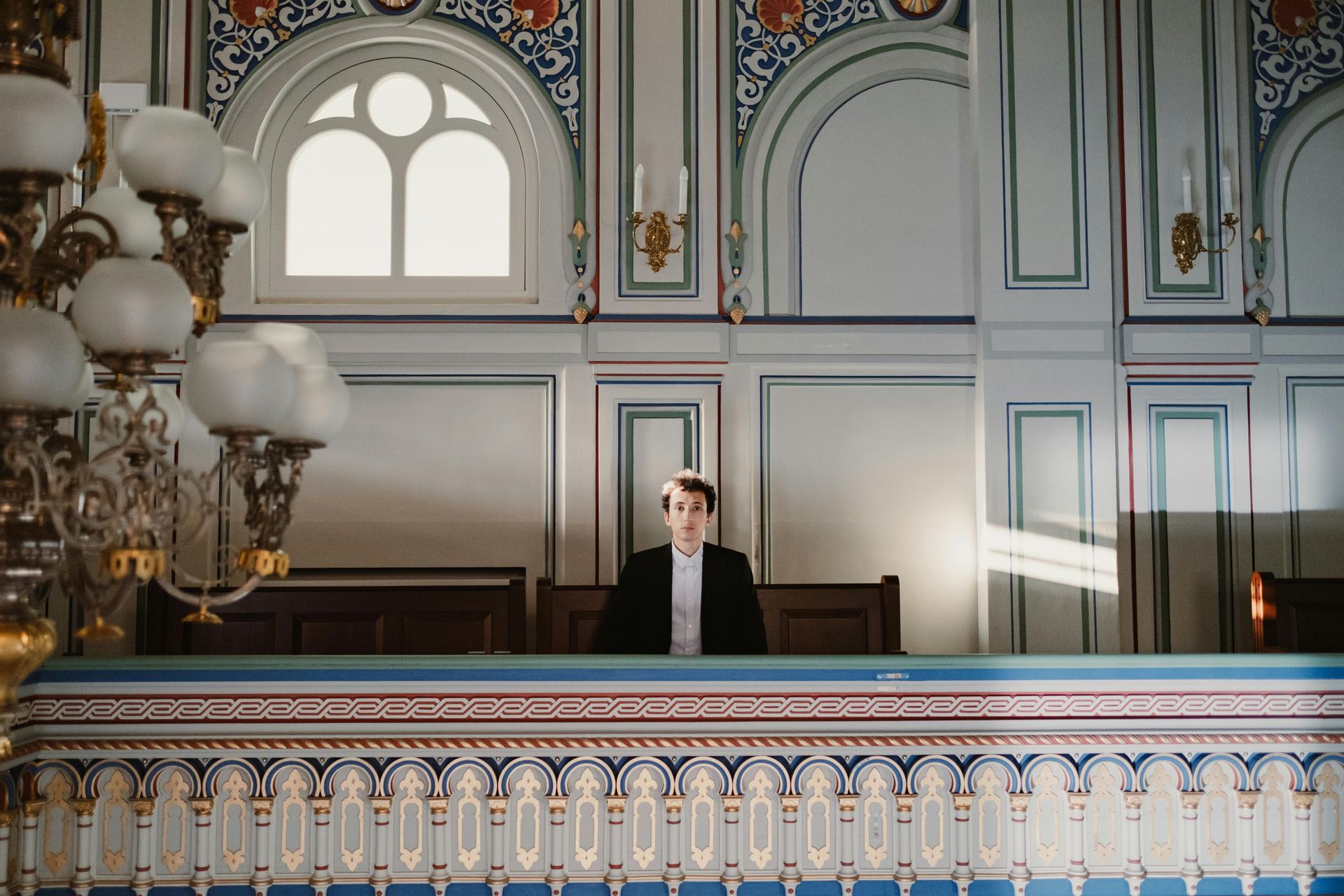 A person in a dark suit sits in an ornate, blue-trimmed balcony area of a building with a large chandelier.