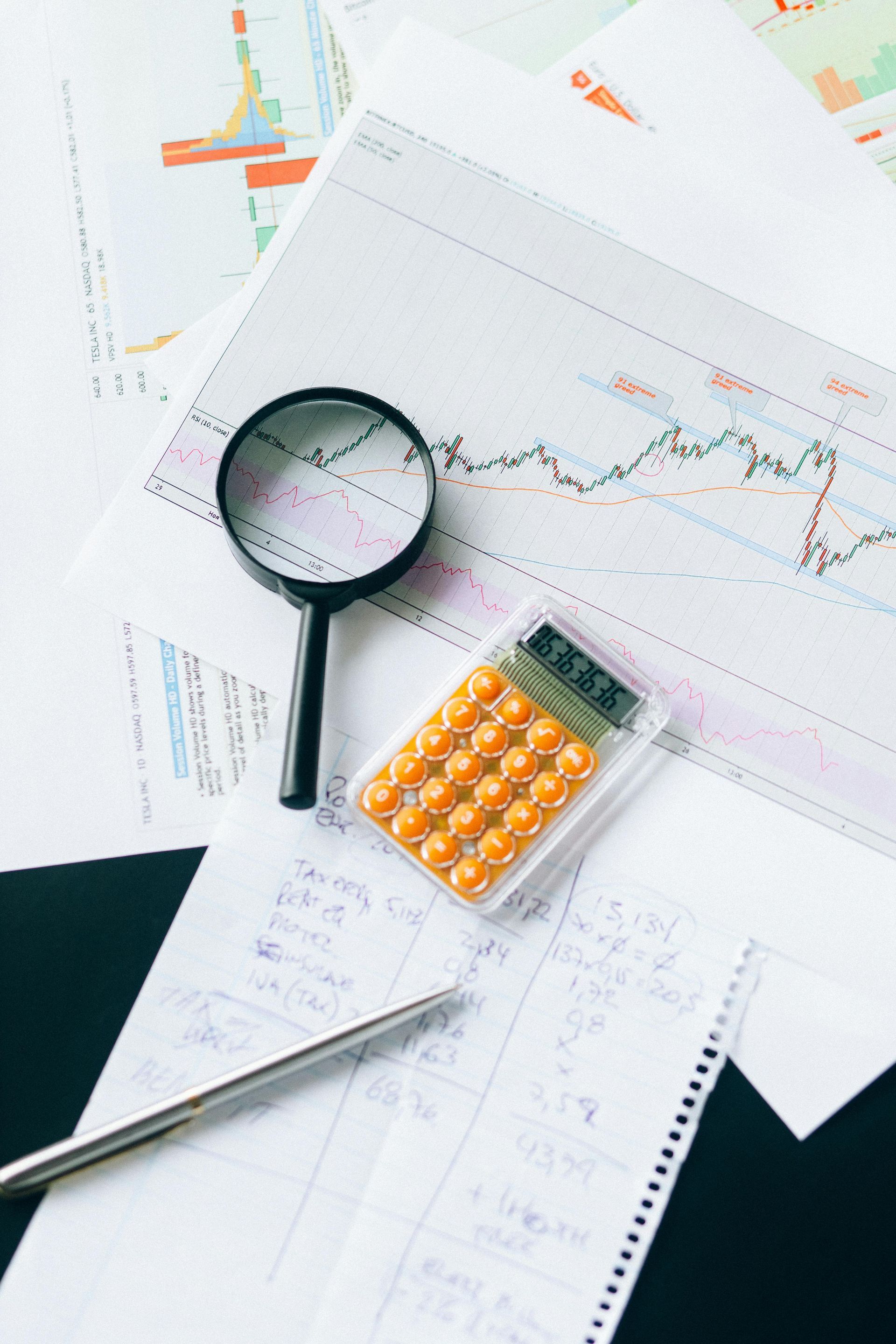 Magnifying glass, calculator, and pen on top of financial charts and notes.