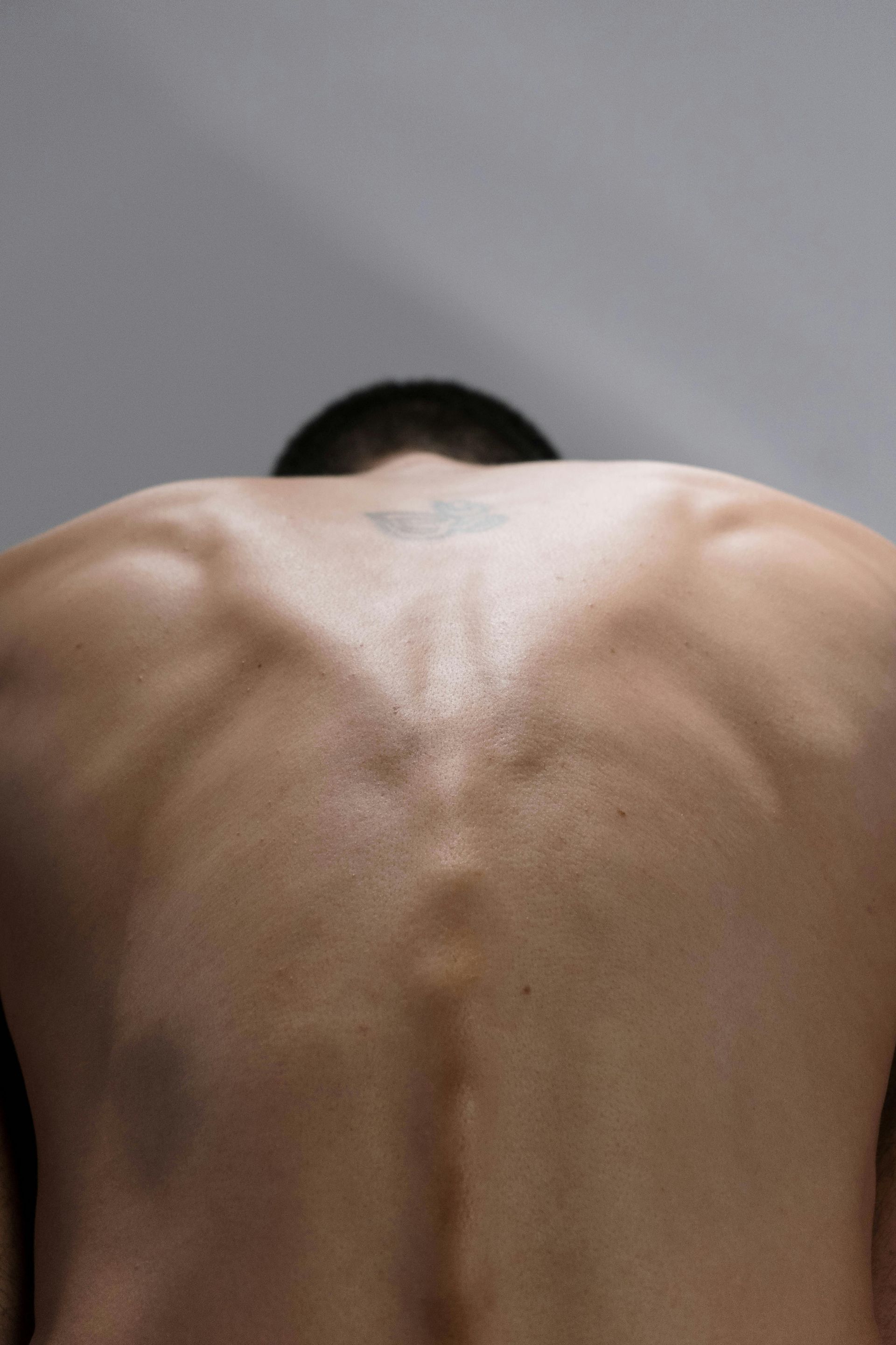 Bare back with defined muscles, slightly bent forward; tattoo visible on the upper back.