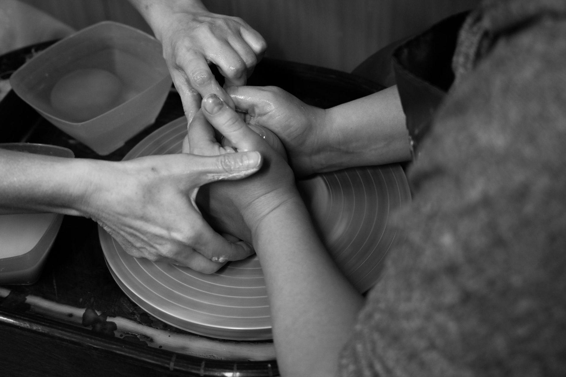 A personâs hands guide another's while shaping wet clay on a pottery wheel.