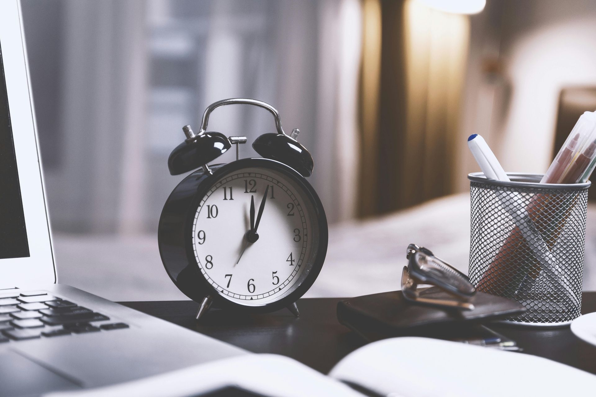 Black alarm clock on a desk with a laptop, pen holder, and open notebook.
