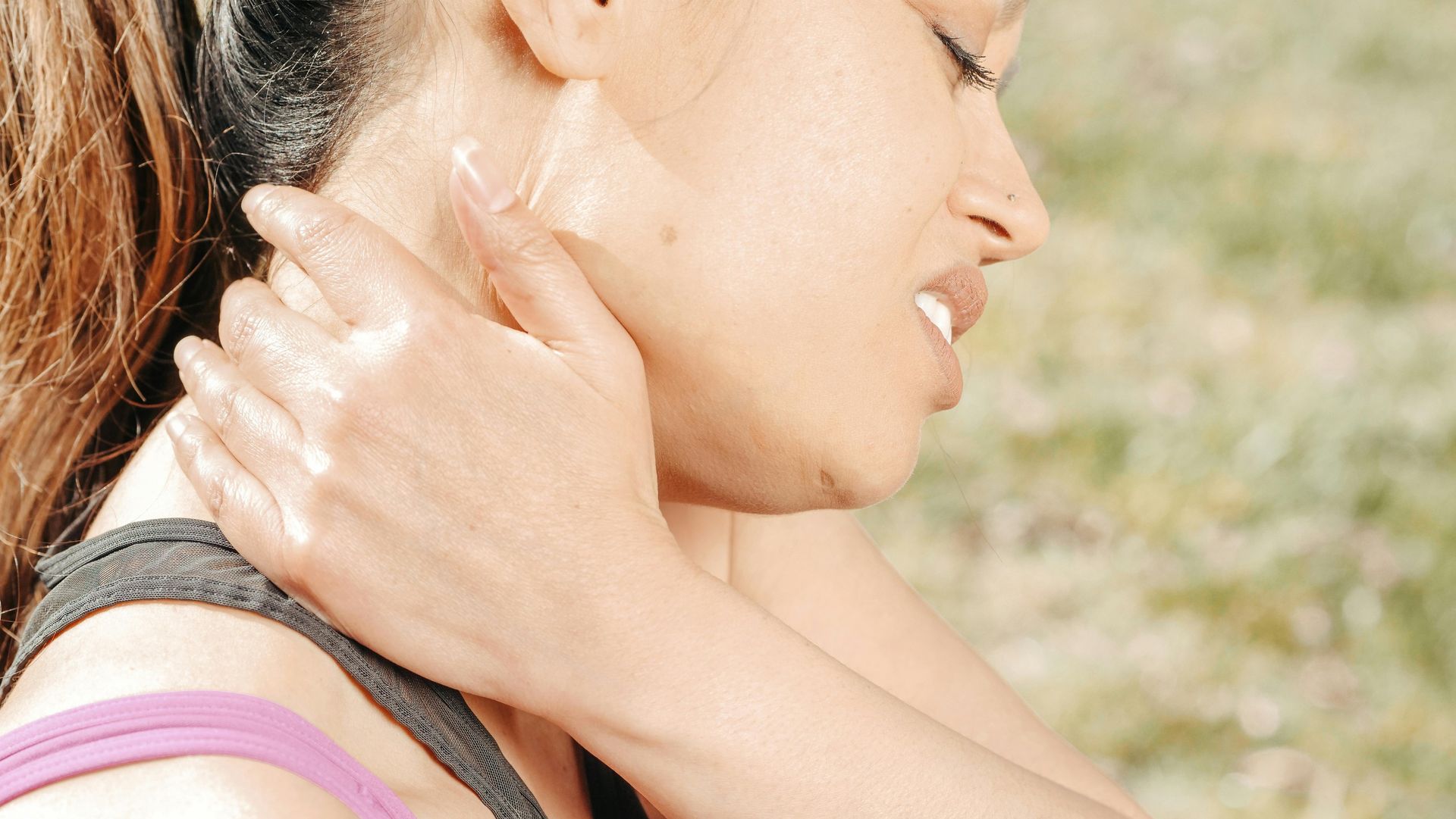 Woman holding neck, eyes closed, sweating, in an outdoor setting.