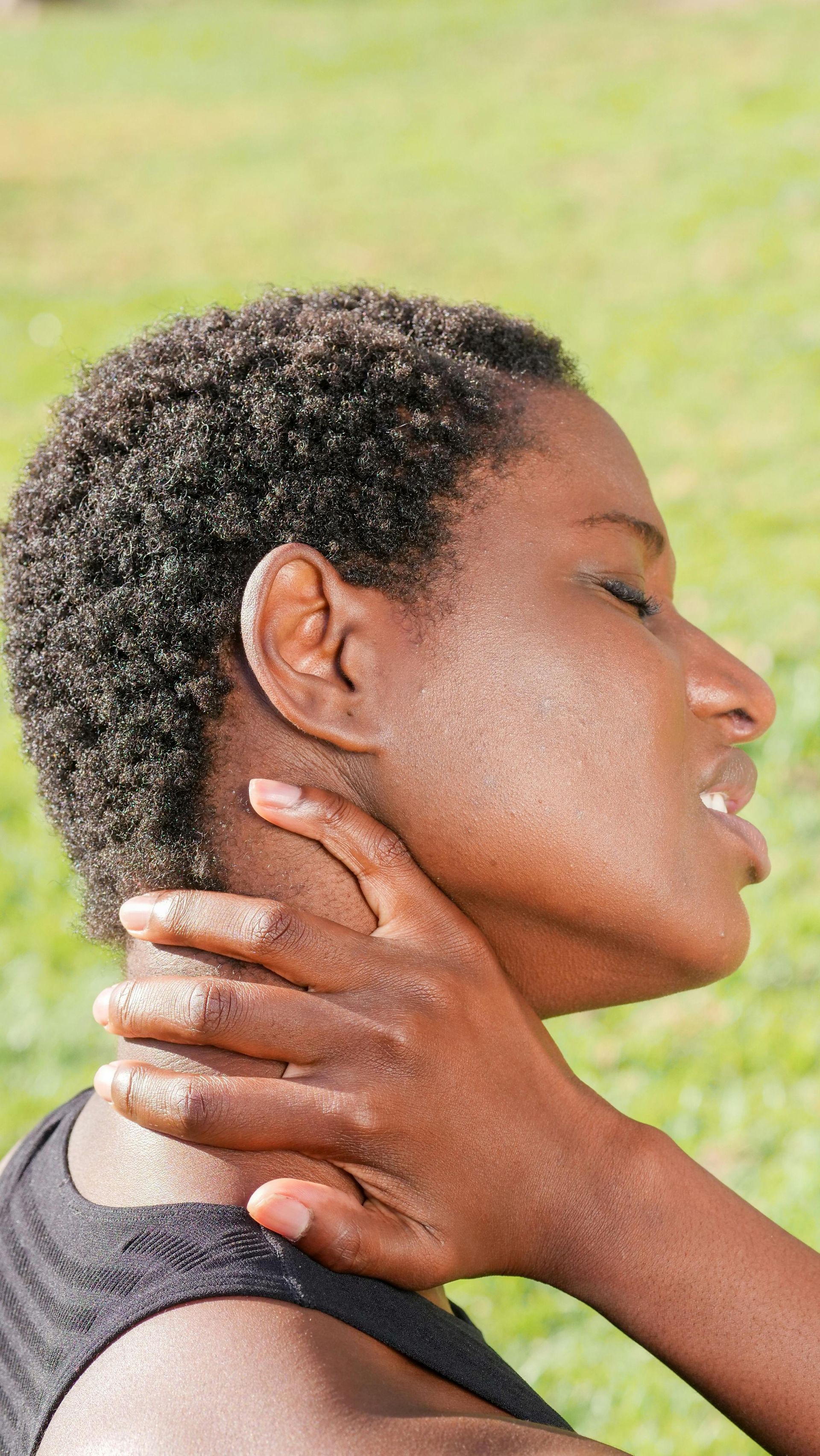 Person clutching the side of their neck with eyes closed against a blurred outdoor background