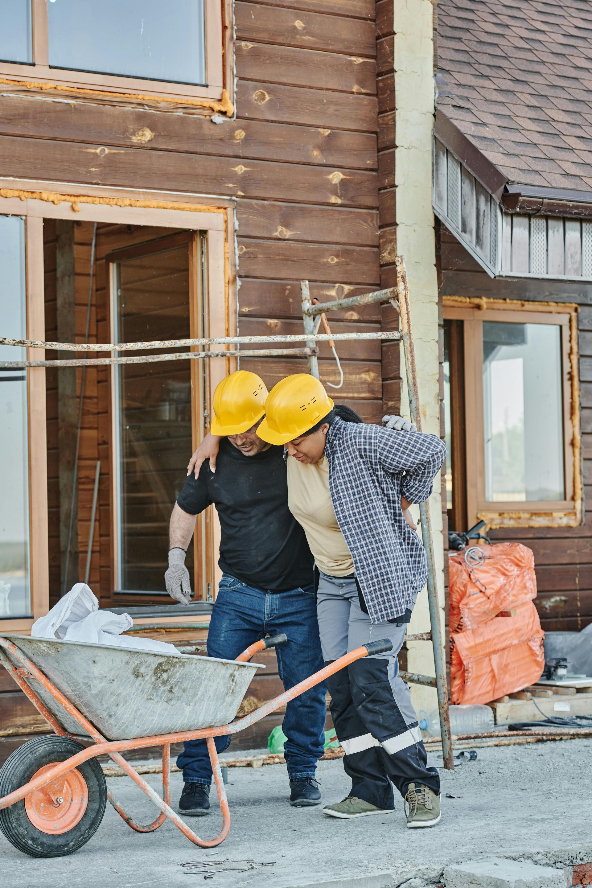 Construction worker injured, being helped by another. They are wearing hard hats near a house under construction.