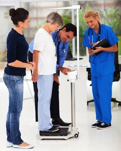 Doctor weighing a patient; a nurse and another person observe in a clinic.