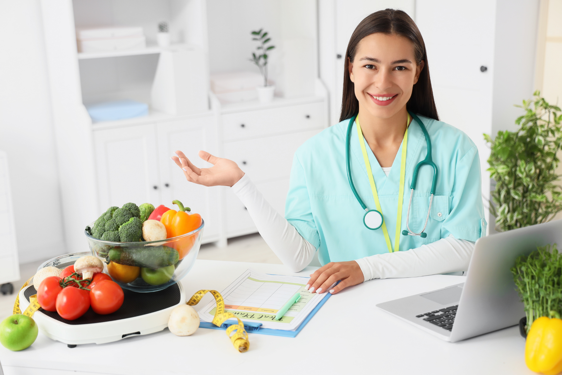 Dietician in scrubs with vegetables, stethoscope, and laptop at desk, smiling.
