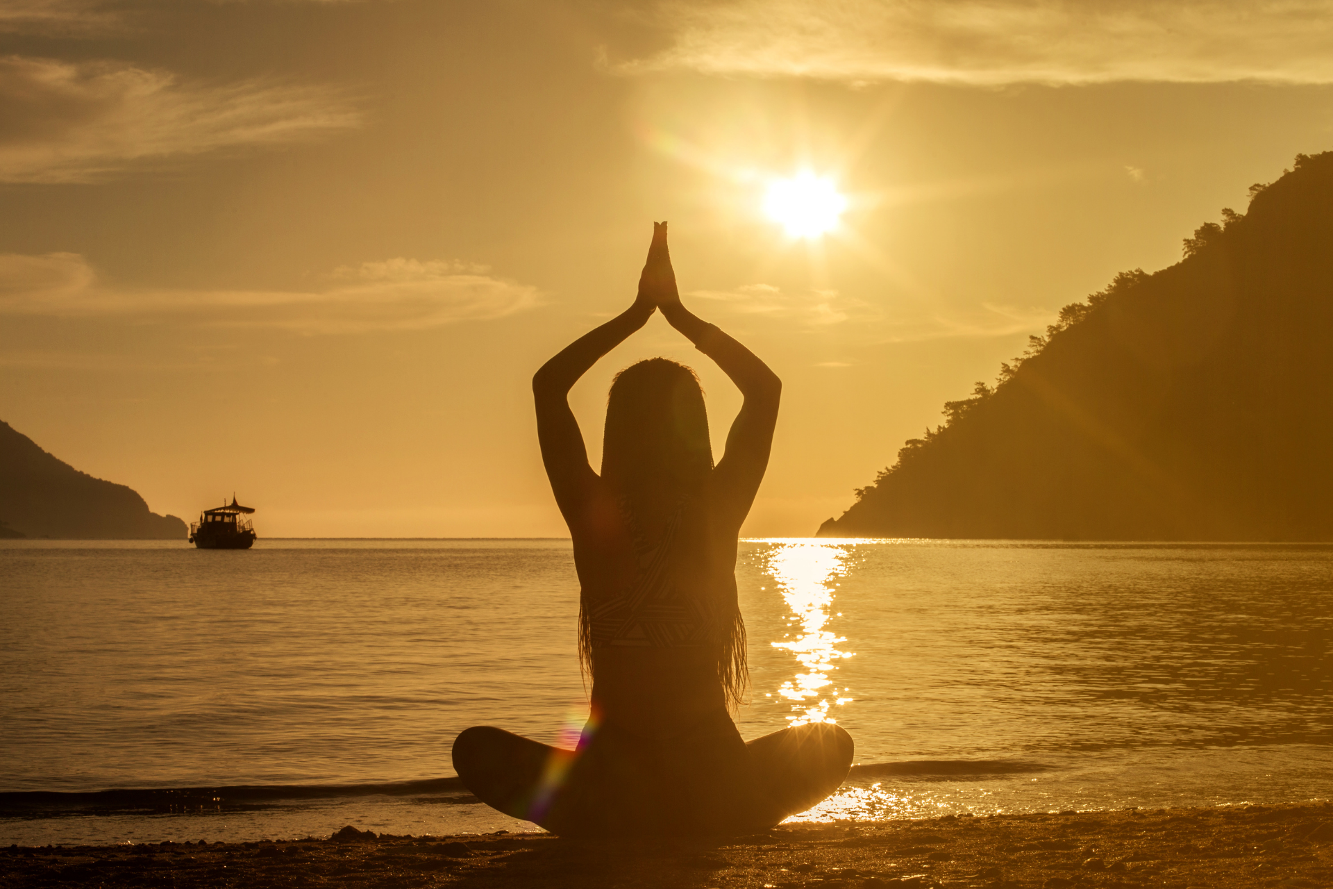 Silhouette of person meditating with hands raised, sun over water.