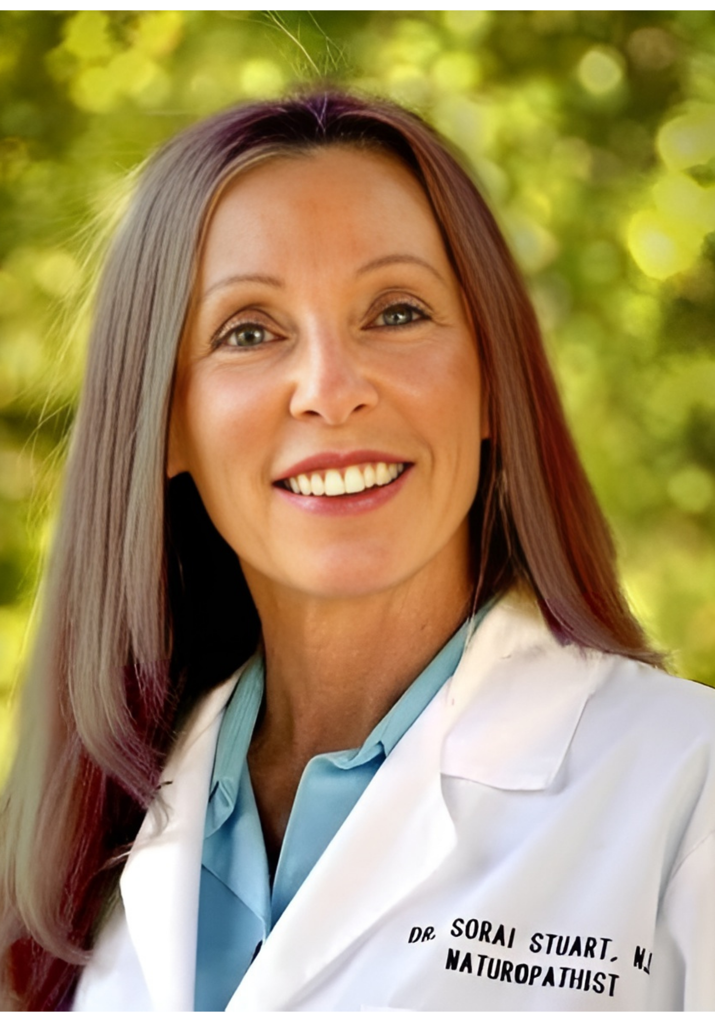 Woman with long hair in white lab coat smiles at the camera, blurred green background.
