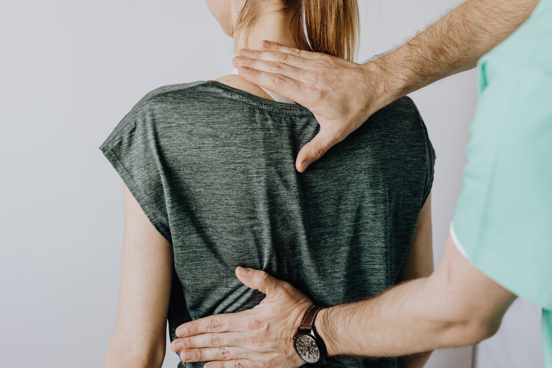 A person's back being examined by a healthcare provider. Hands touching back, white wall background.