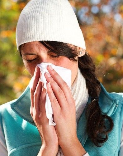 Woman wearing a white hat and scarf, blowing her nose with a tissue outdoors.