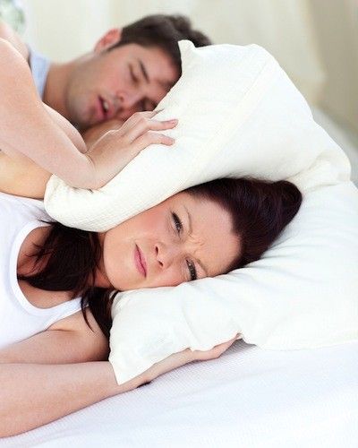 Woman annoyed, covering ears with pillow while man sleeps and snores in bed.