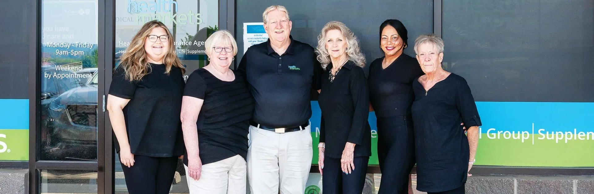 A group of seven people pose outside a building, all wearing dark tops and various bottoms, smiling.