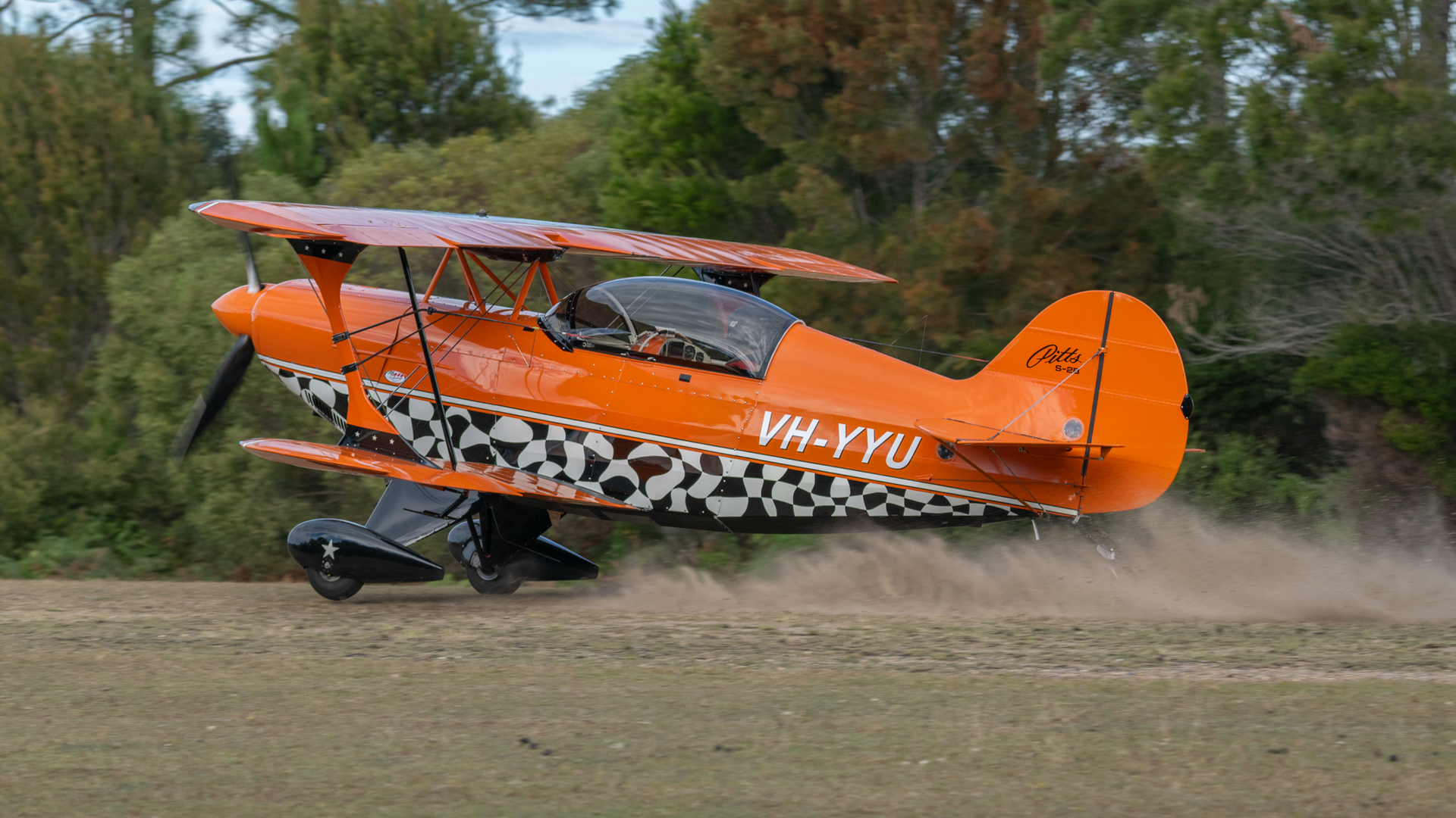 A small orange airplane is taking off from a dirt field.