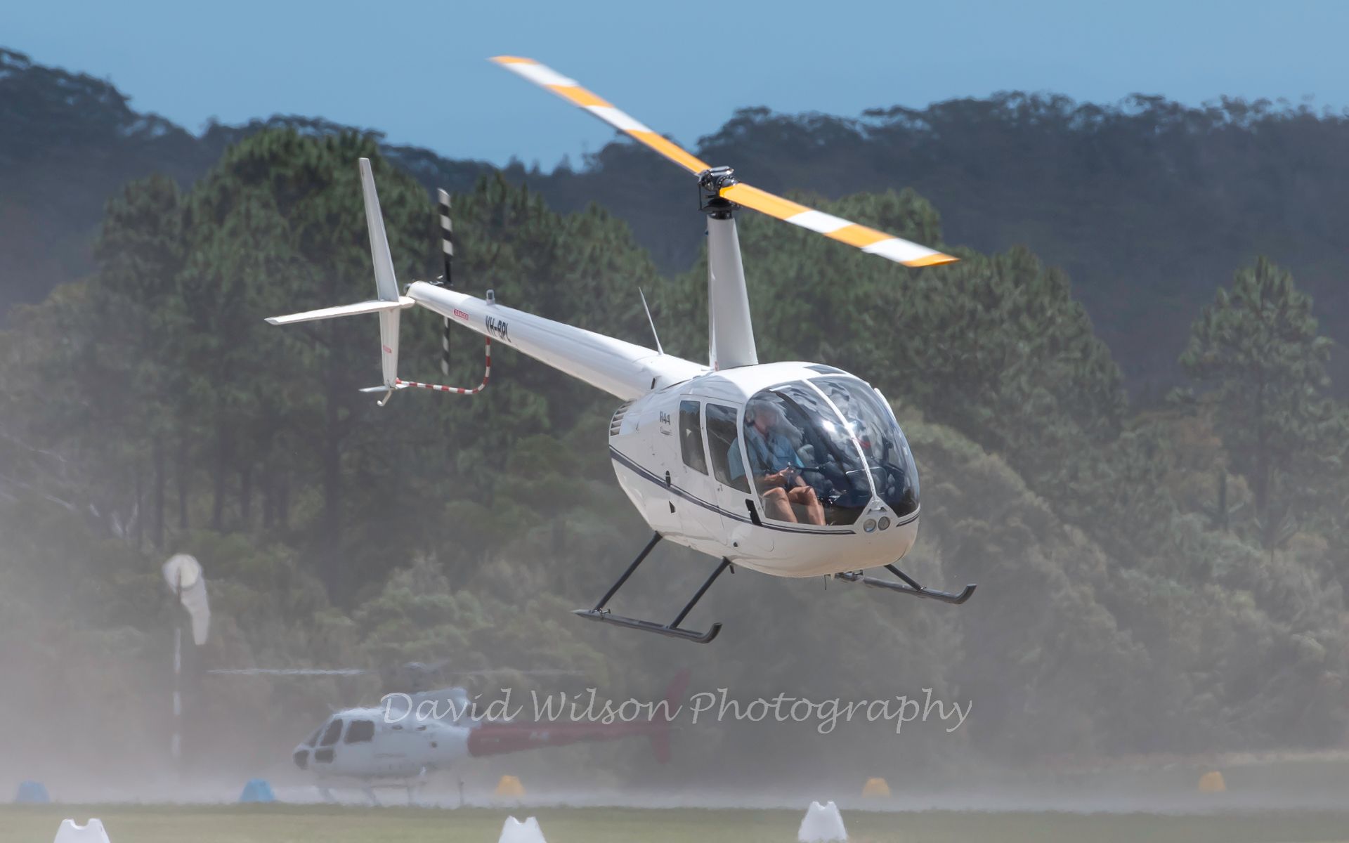 a white helicopter is flying over a field with trees in the background