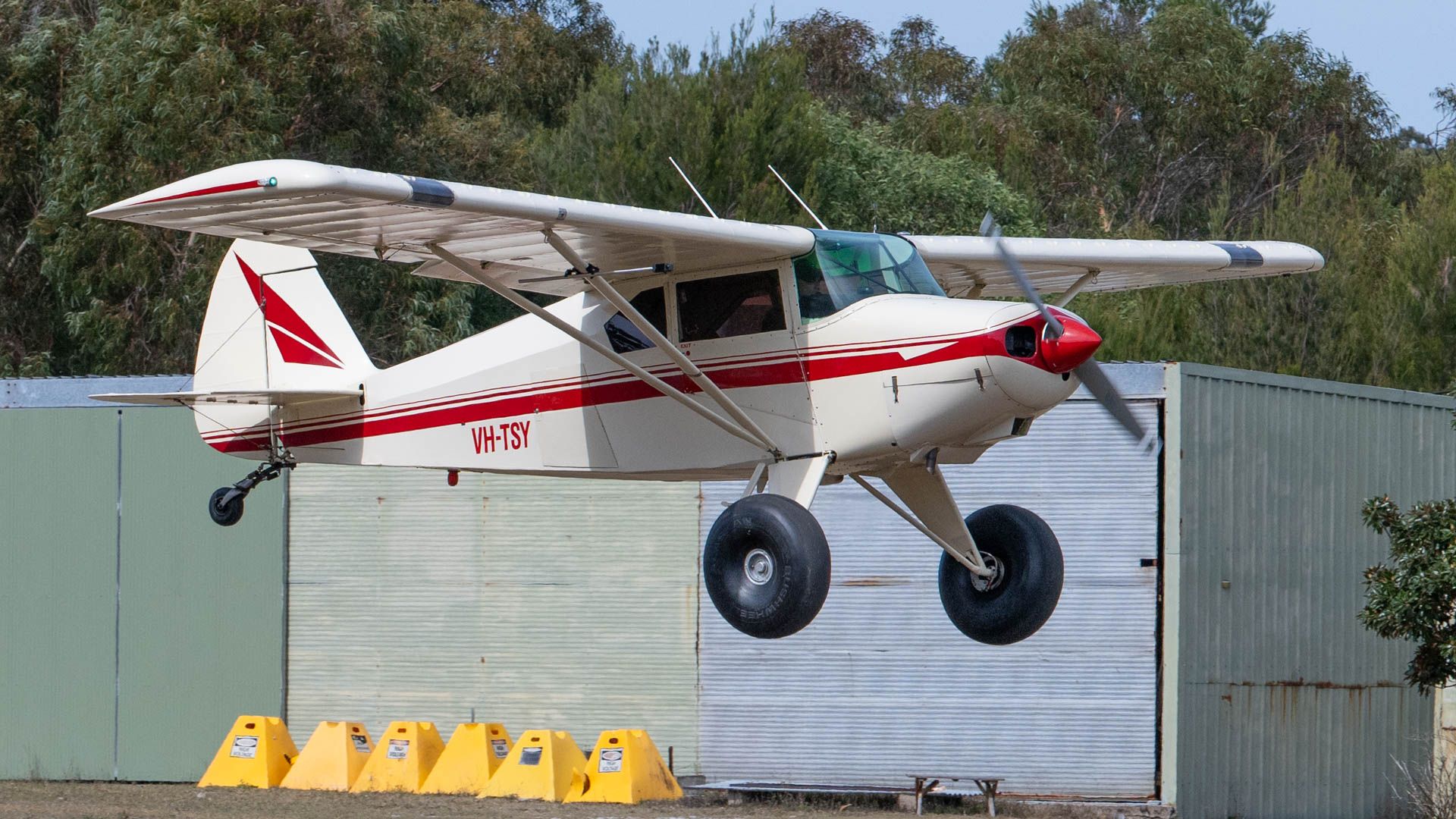 White and red bush plane taking off near a green hangar.
