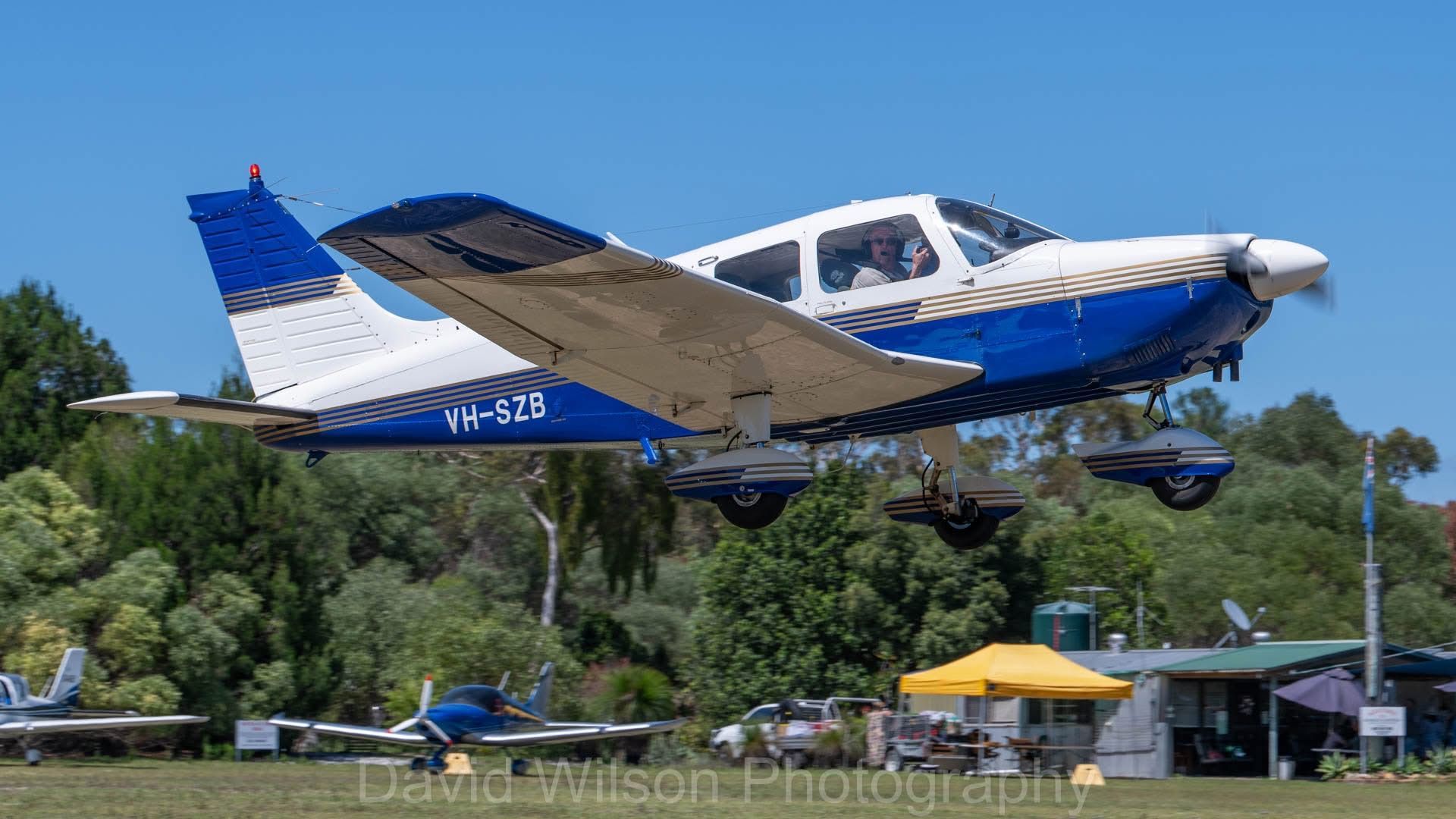 Blue and white airplane taking off over grassy field and buildings on a sunny day.