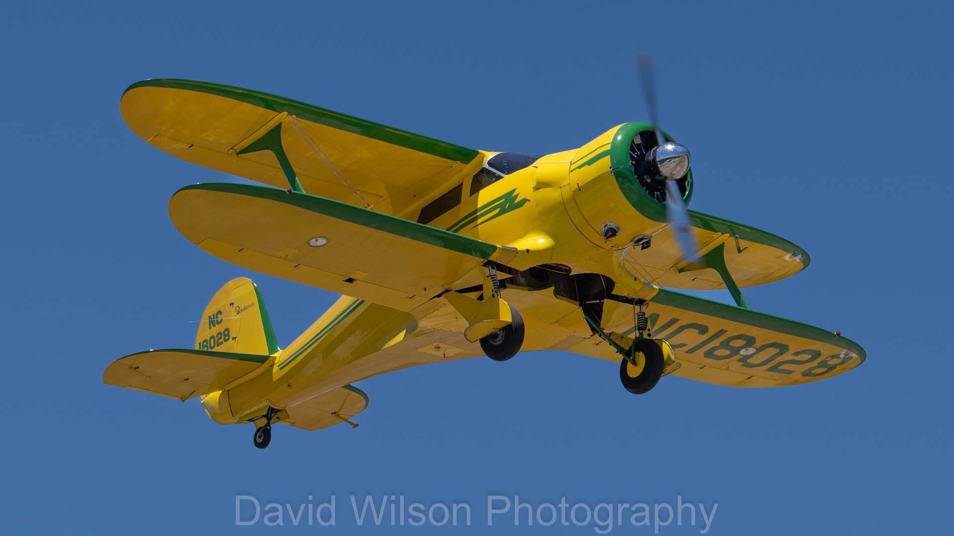 Yellow and green biplane in flight against a clear blue sky.