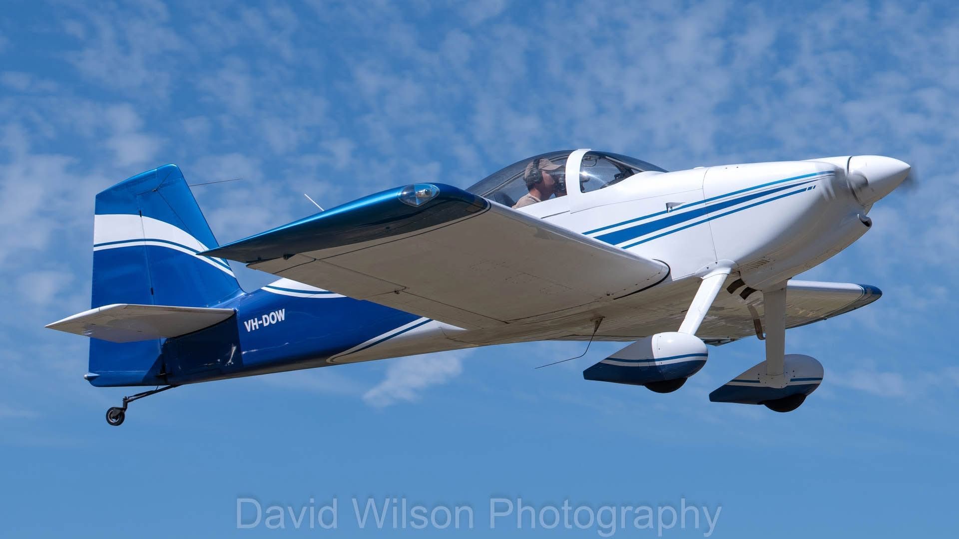 White and blue single-engine airplane in flight against a blue sky, displaying a propeller and landing gear.