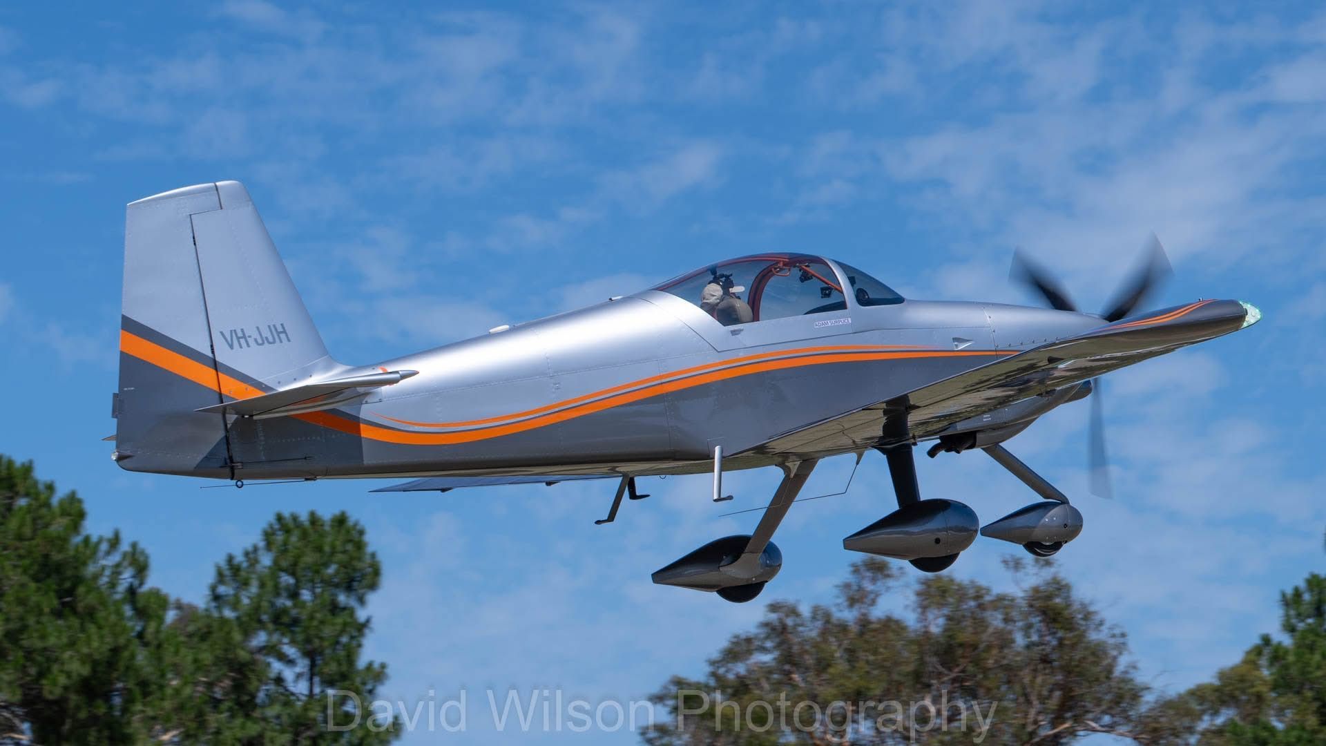 Silver and orange single-engine airplane in flight against a blue sky, trees in the background.