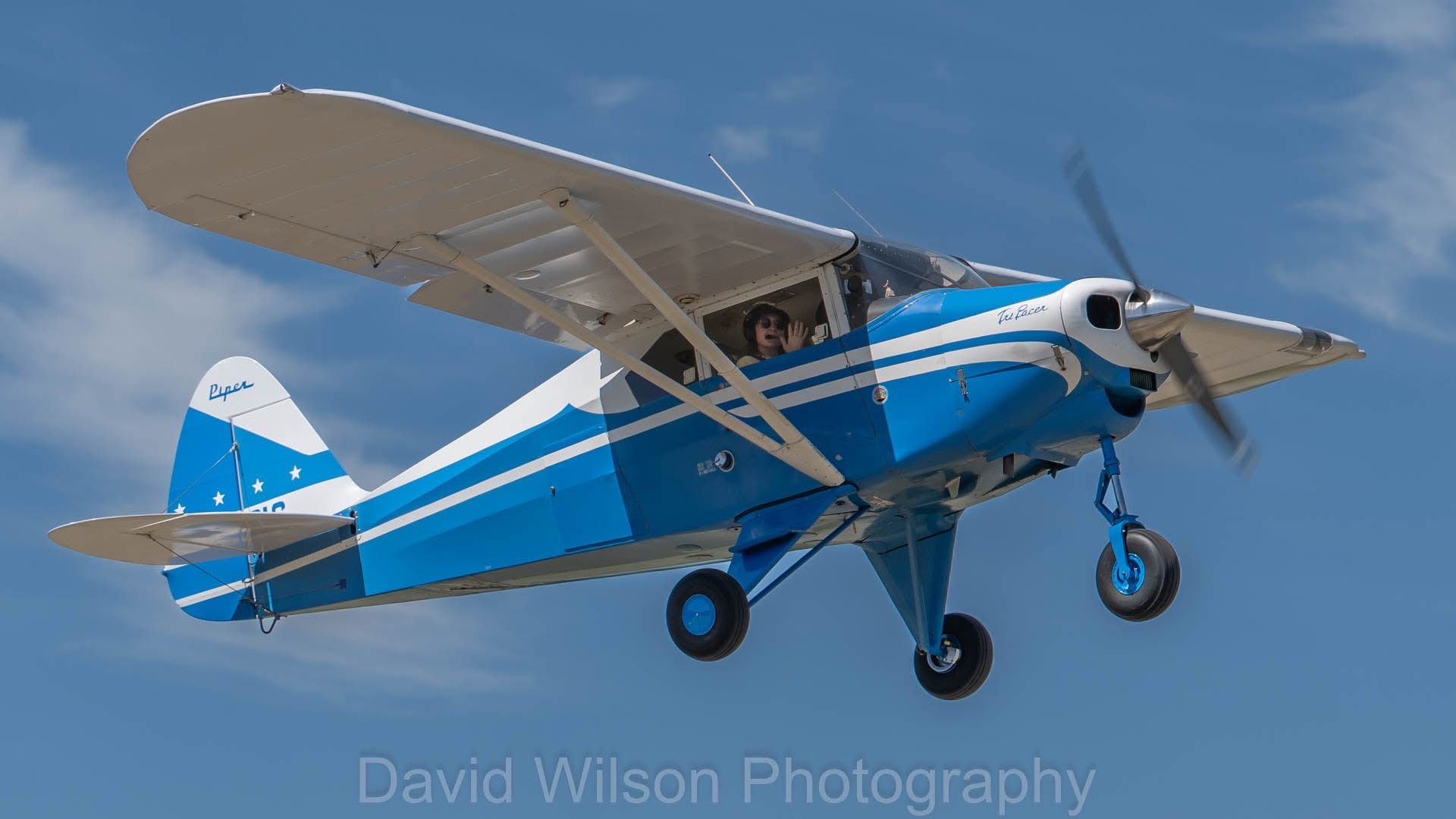 Blue and white single-engine aircraft in flight, propeller spinning. Pilot visible in cockpit.