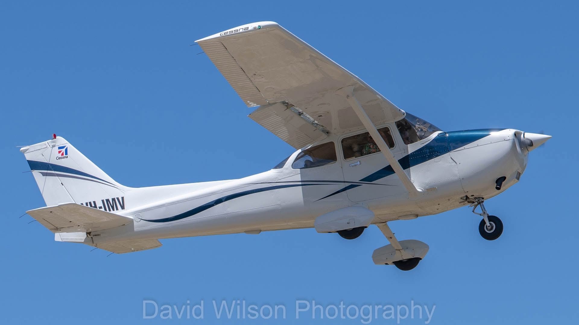 White and blue Cessna airplane in flight against a clear blue sky.