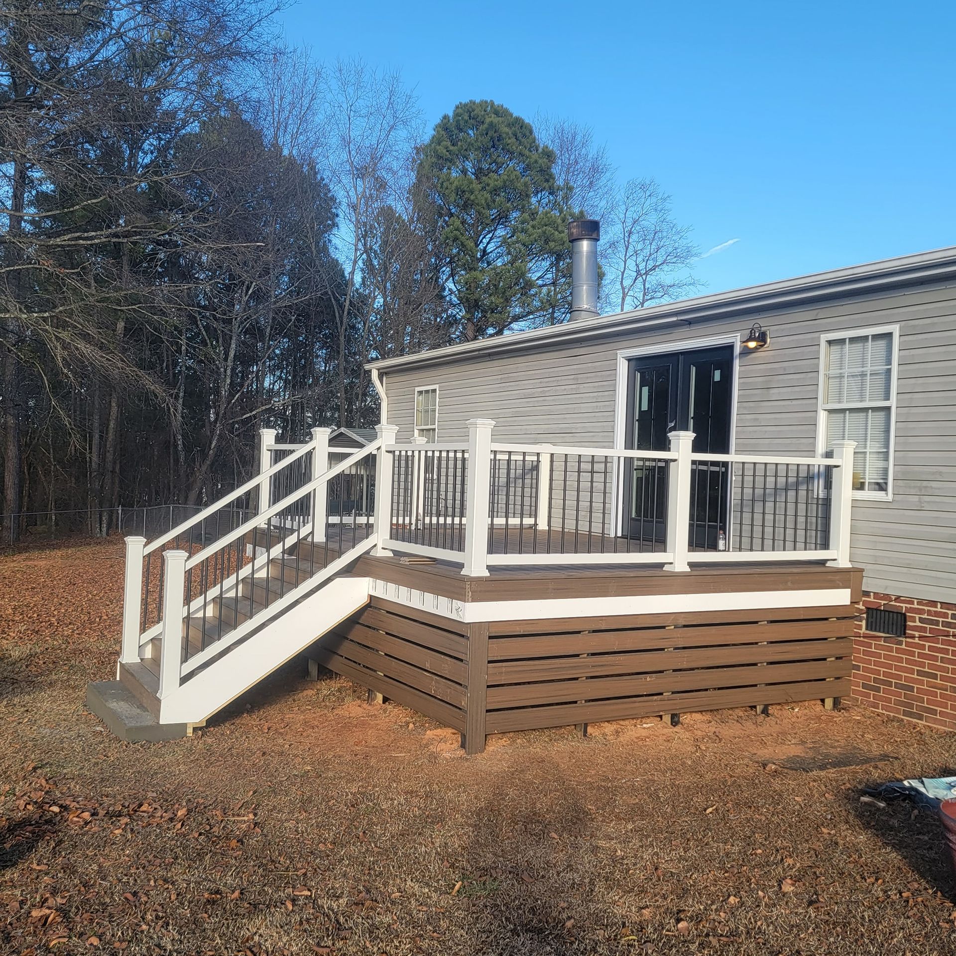 A wooden deck with white railing, stairs, and dark balusters, attached to a gray house, set outside.