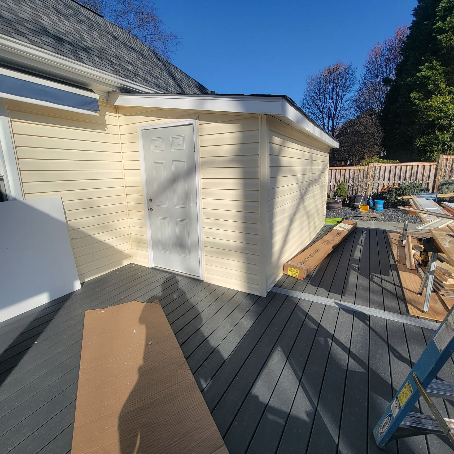 Shed with cream siding and gray door, built on a gray composite deck, with wood and tools.