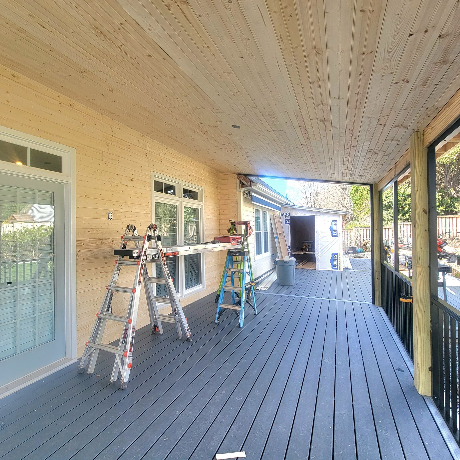 Covered porch under construction: gray deck, wood ceiling, ladders, and tools present.