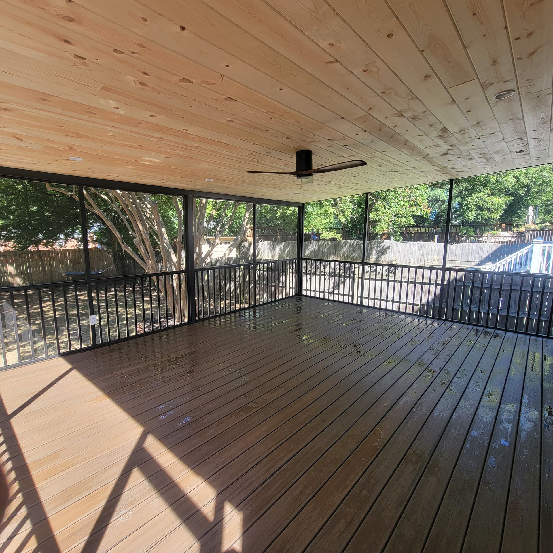 Screened-in porch with wood deck and ceiling; black railings; ceiling fan; sunny outdoor view.