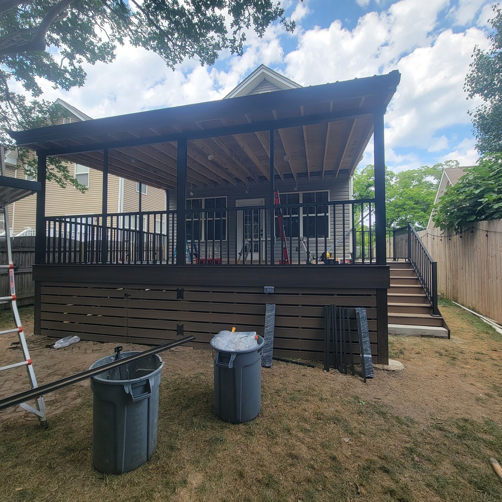 Backyard deck with pergola and stairs, surrounded by a fence and trees. Two trash cans in foreground.