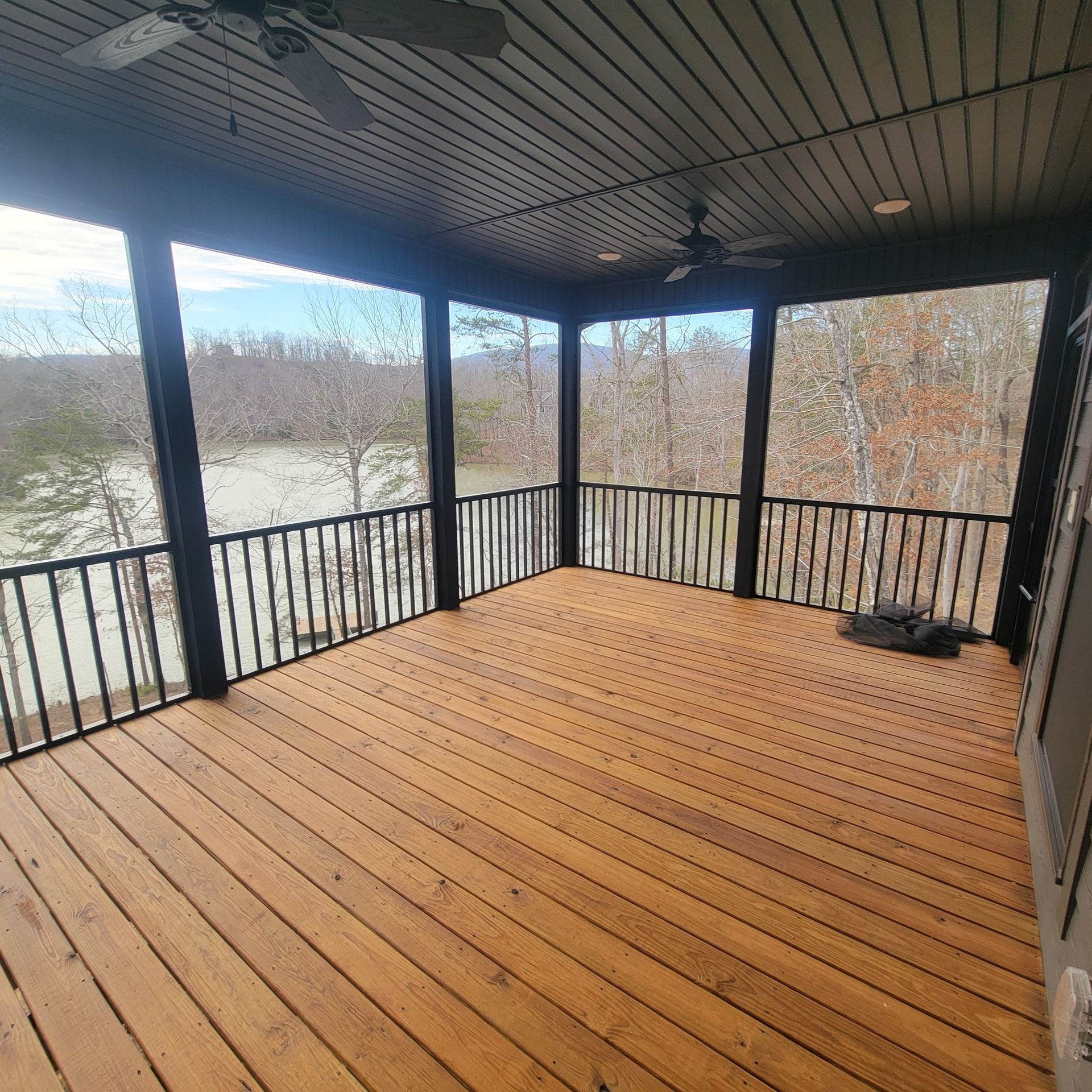 Screened porch with wooden deck, black railings, and ceiling. Overlooks a lake and trees.
