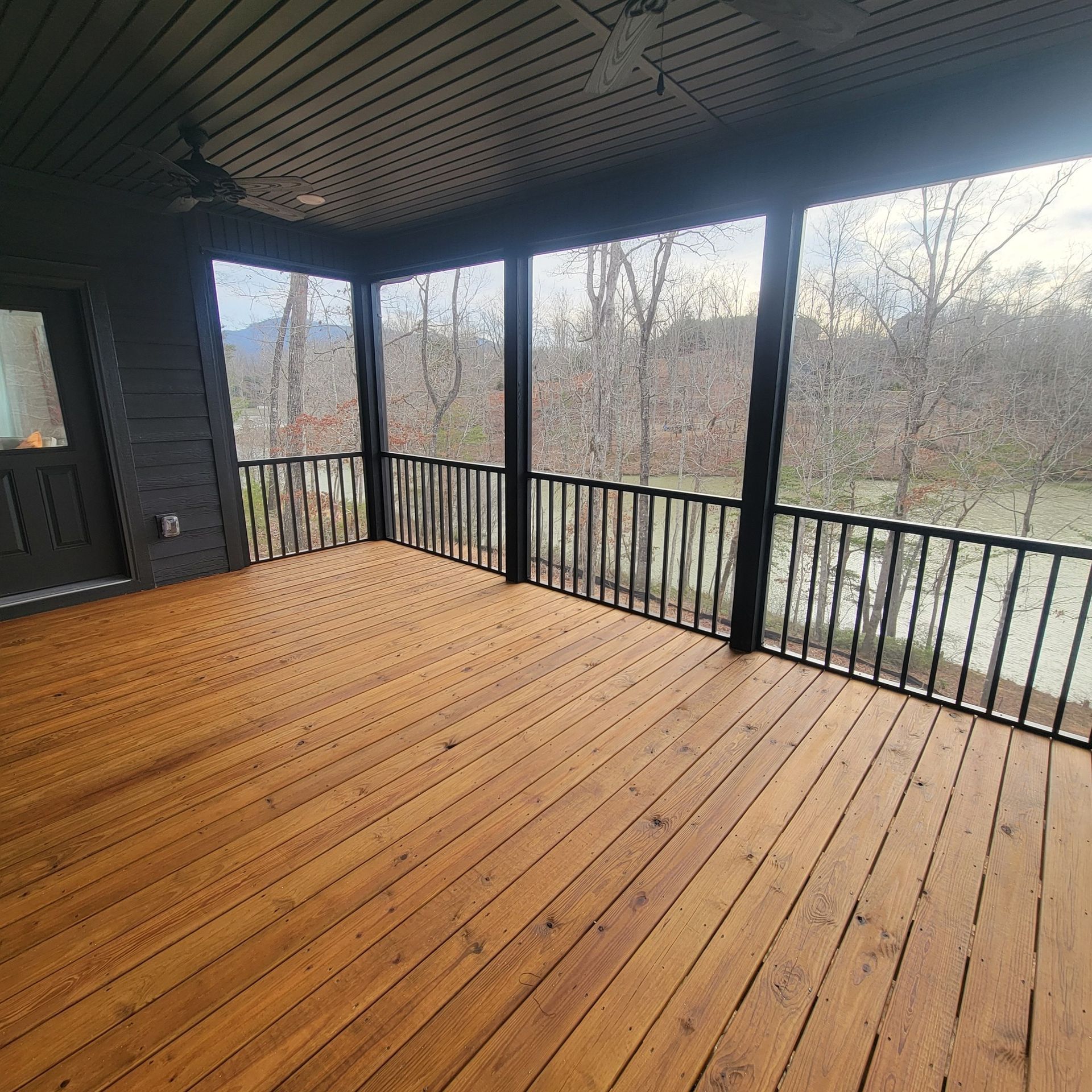 Covered porch with stained wooden floor, black railing, and screens overlooking trees and water.
