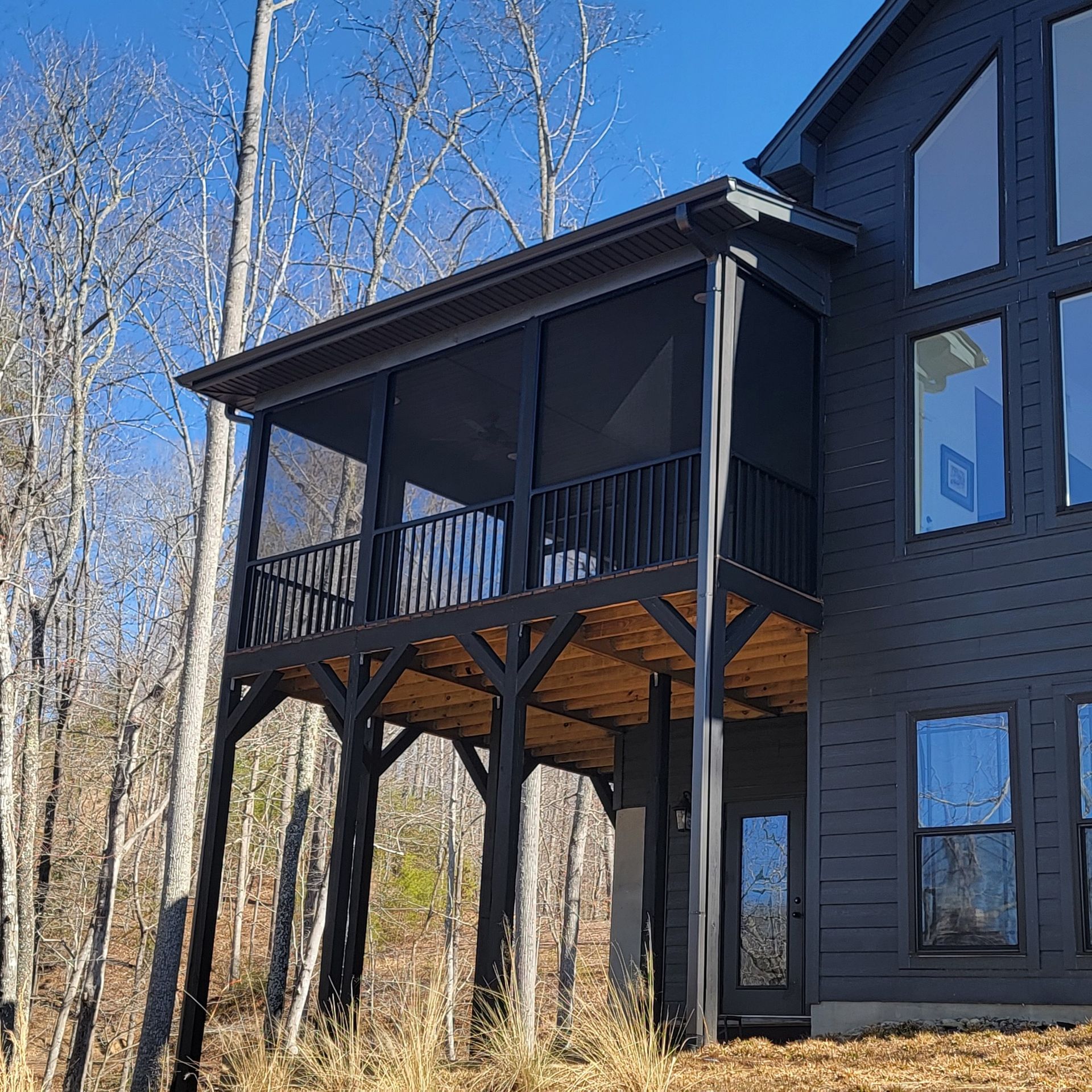 Black screened porch with wooden beams, attached to a dark gray house in a wooded area.