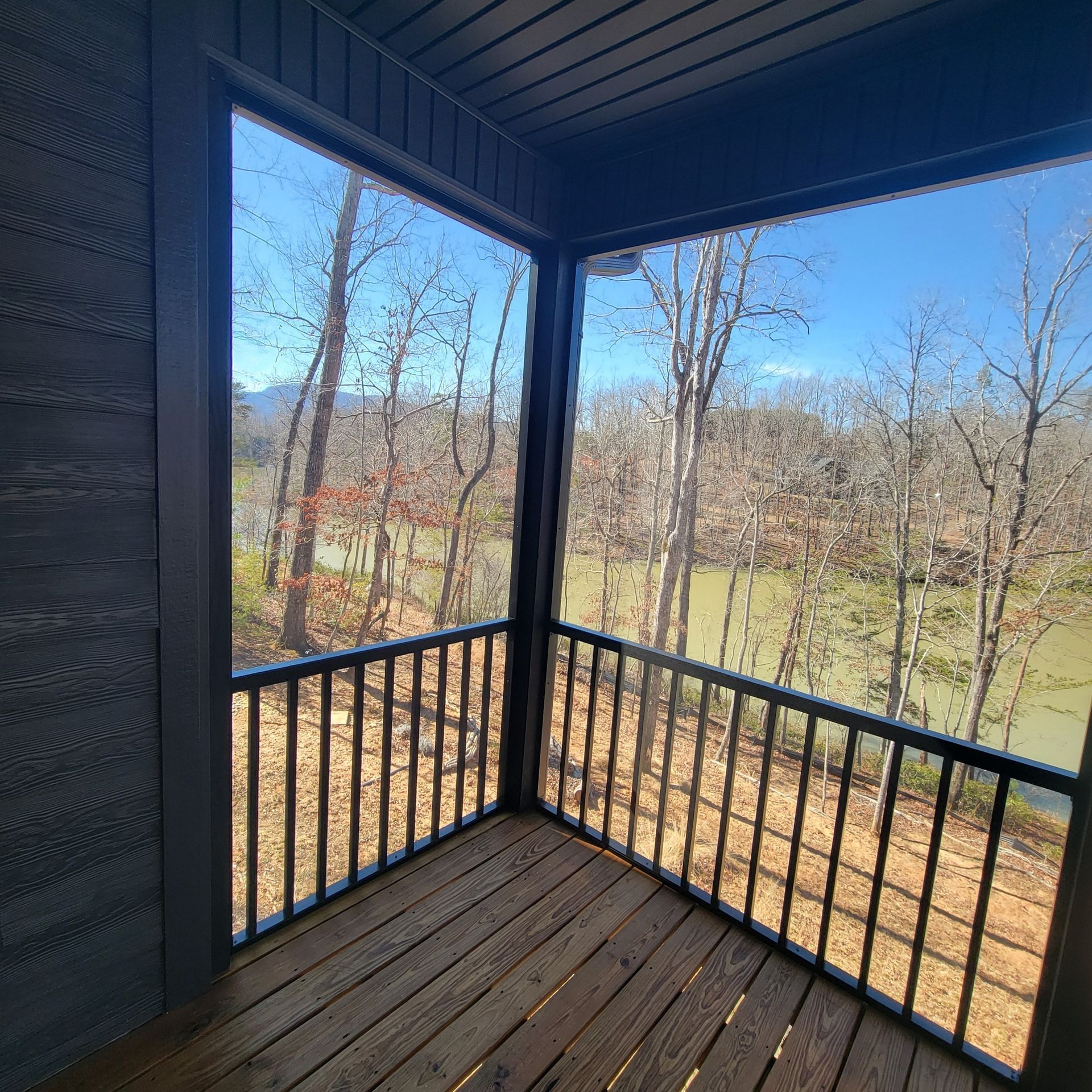 Screened-in porch with wood deck and black railing overlooking a river and trees under a blue sky.