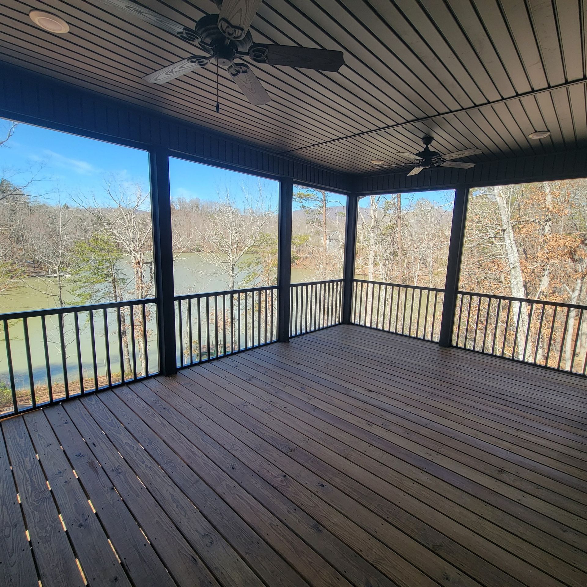 Covered wooden deck overlooking a lake, framed by black railings and columns. Two ceiling fans.