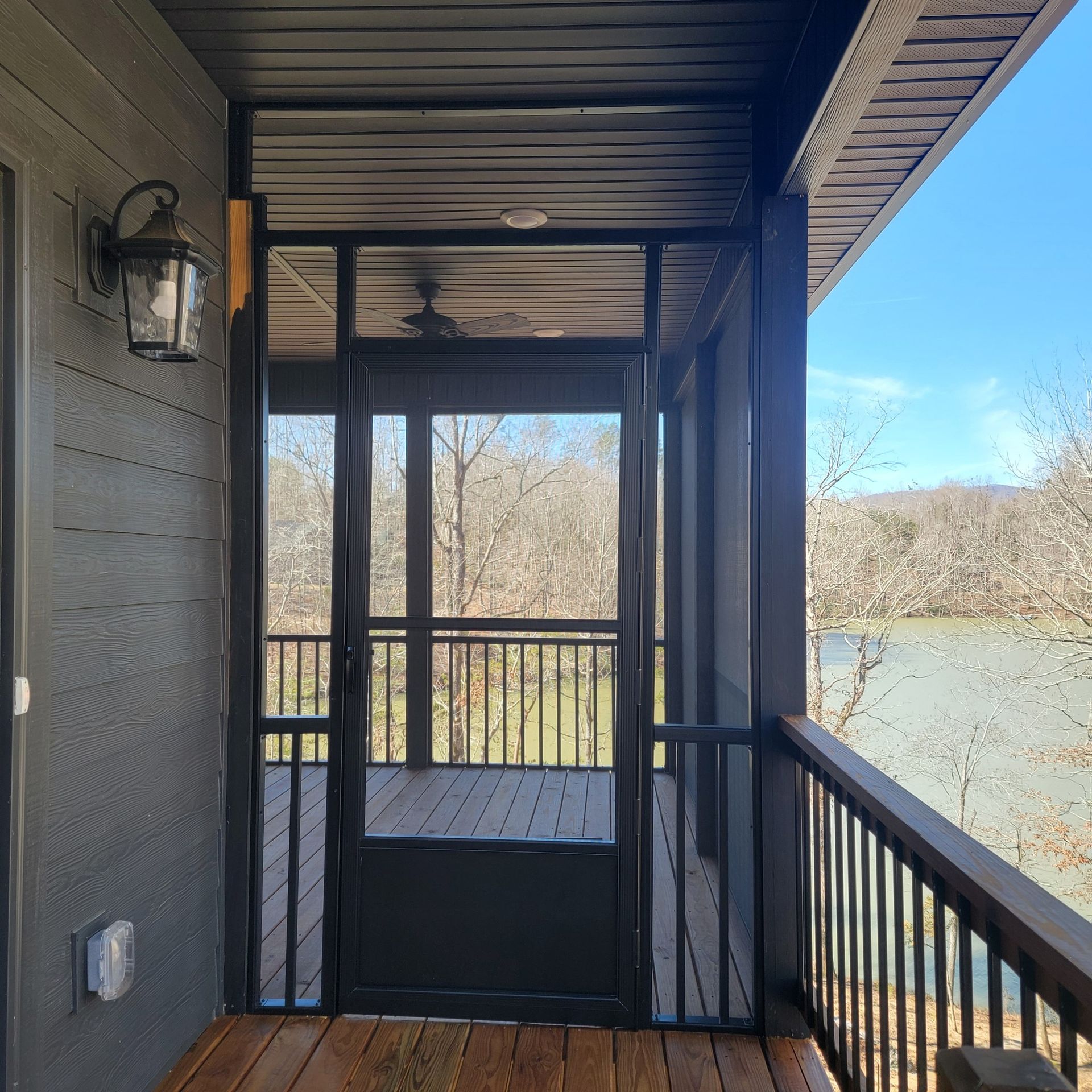Screened-in porch with black frames, wooden deck, railing, and view of trees and water.