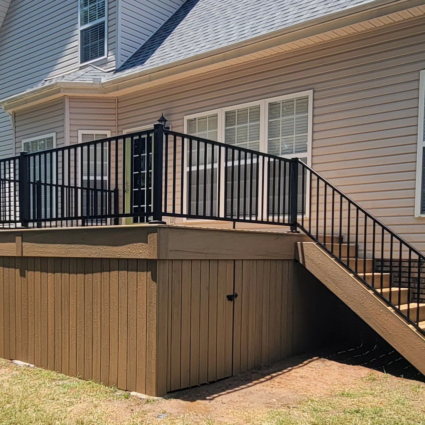 Deck with black railing, brown skirting, steps, and door against a beige house with windows.