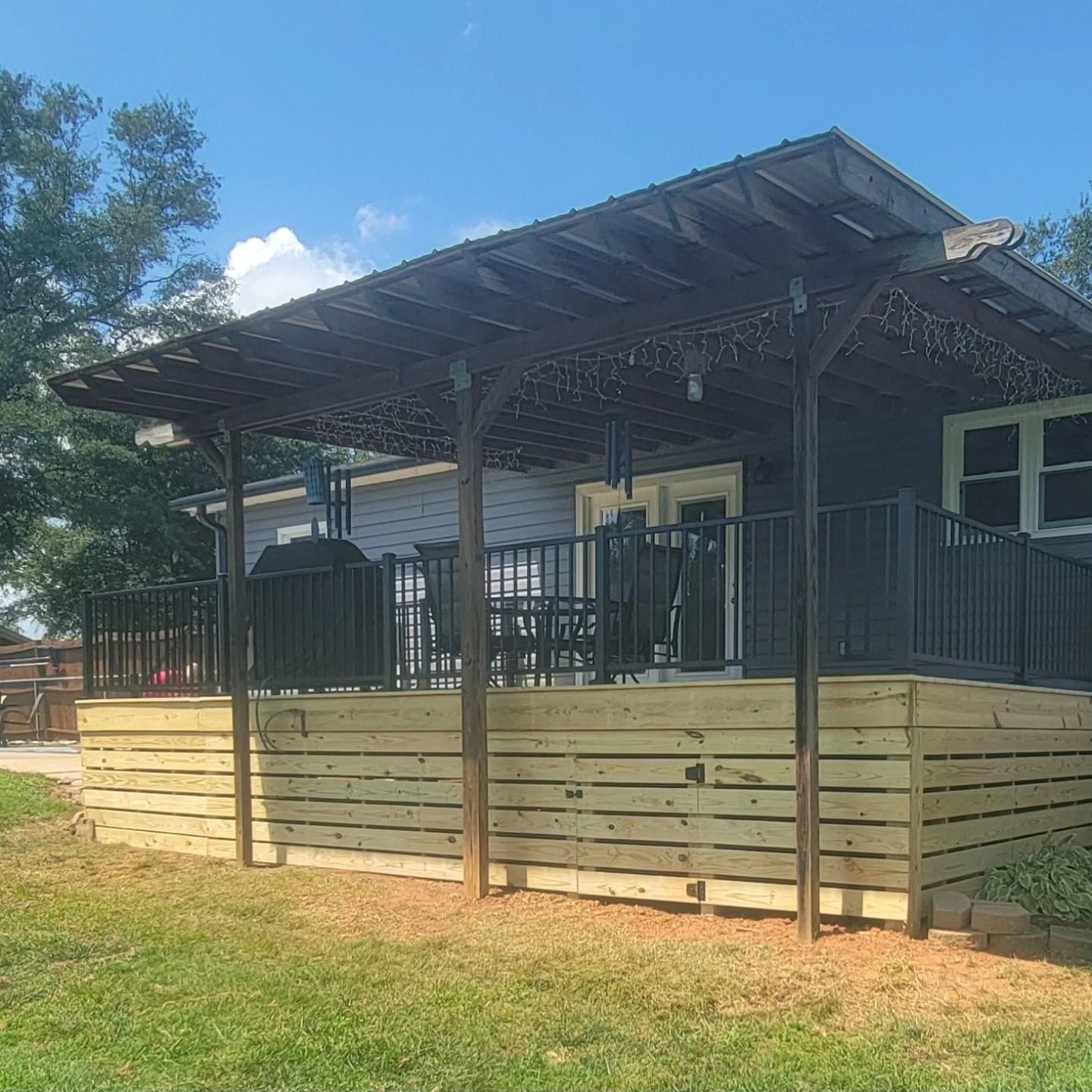 Wooden deck with a covered patio, black grill and furniture, next to a blue house on a sunny day.