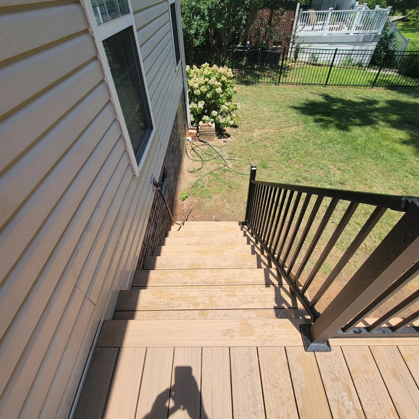 Wooden deck with stairs, black railing, descending to a grassy yard with greenery and a white fence.