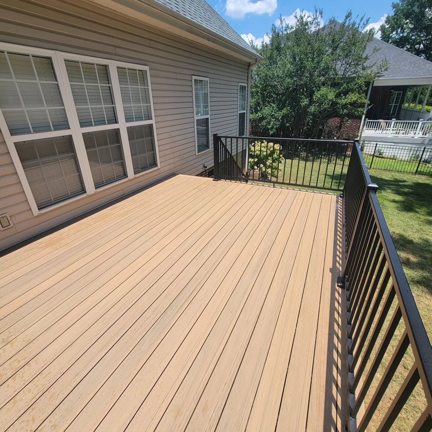 Wooden deck attached to a light-colored house with black railings, overlooking a grassy yard under a blue sky.